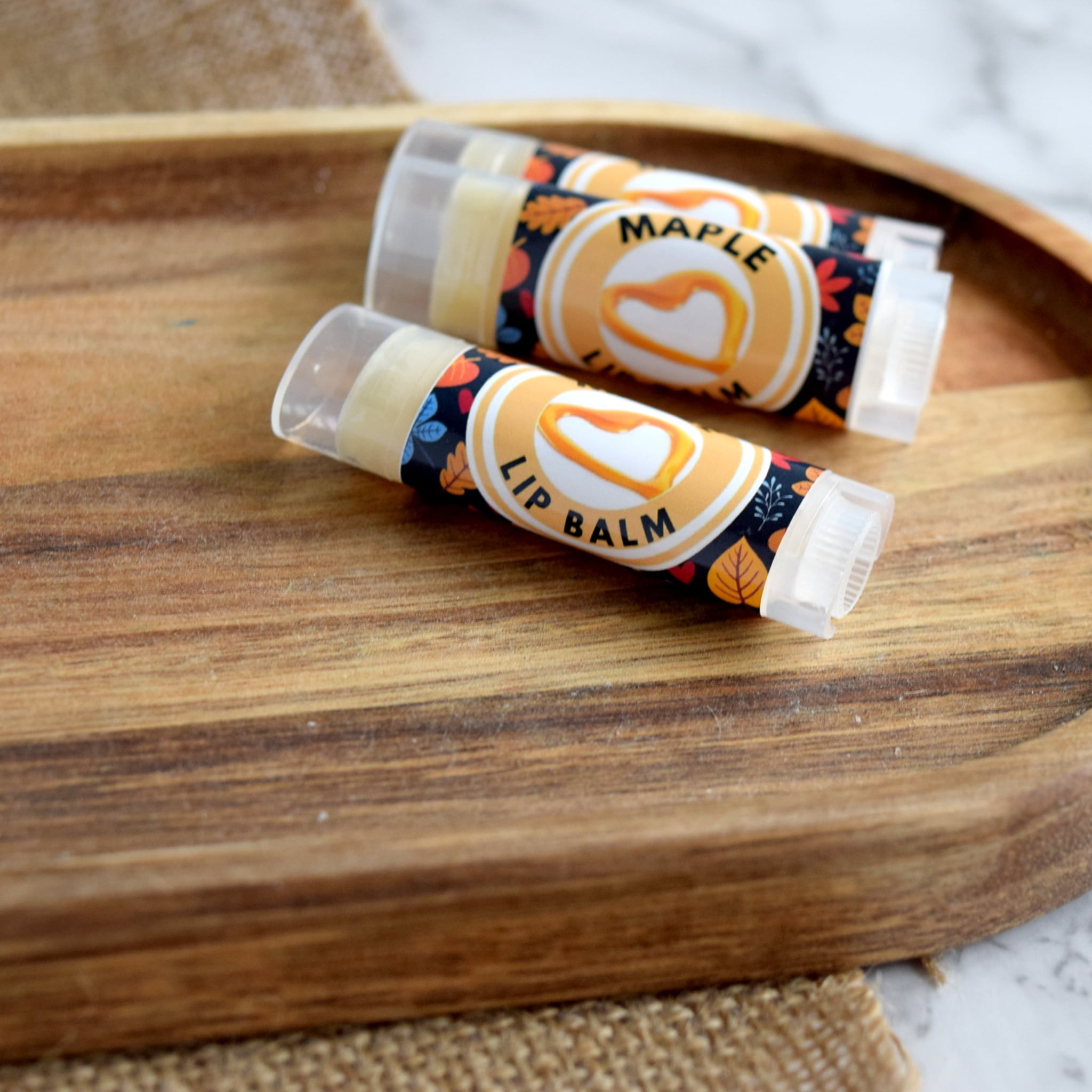 Lip balm tubes with orange and black label, fall leaf pattern, and clear plastic cap on wood tray