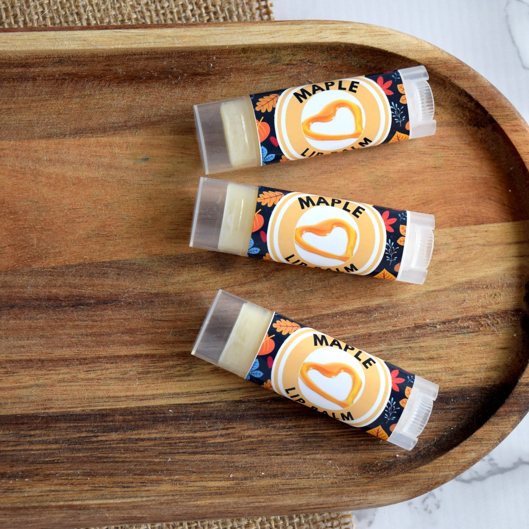 Lip balm tubes with "Maple" text, clear plastic, and autumn-themed label on wooden tray