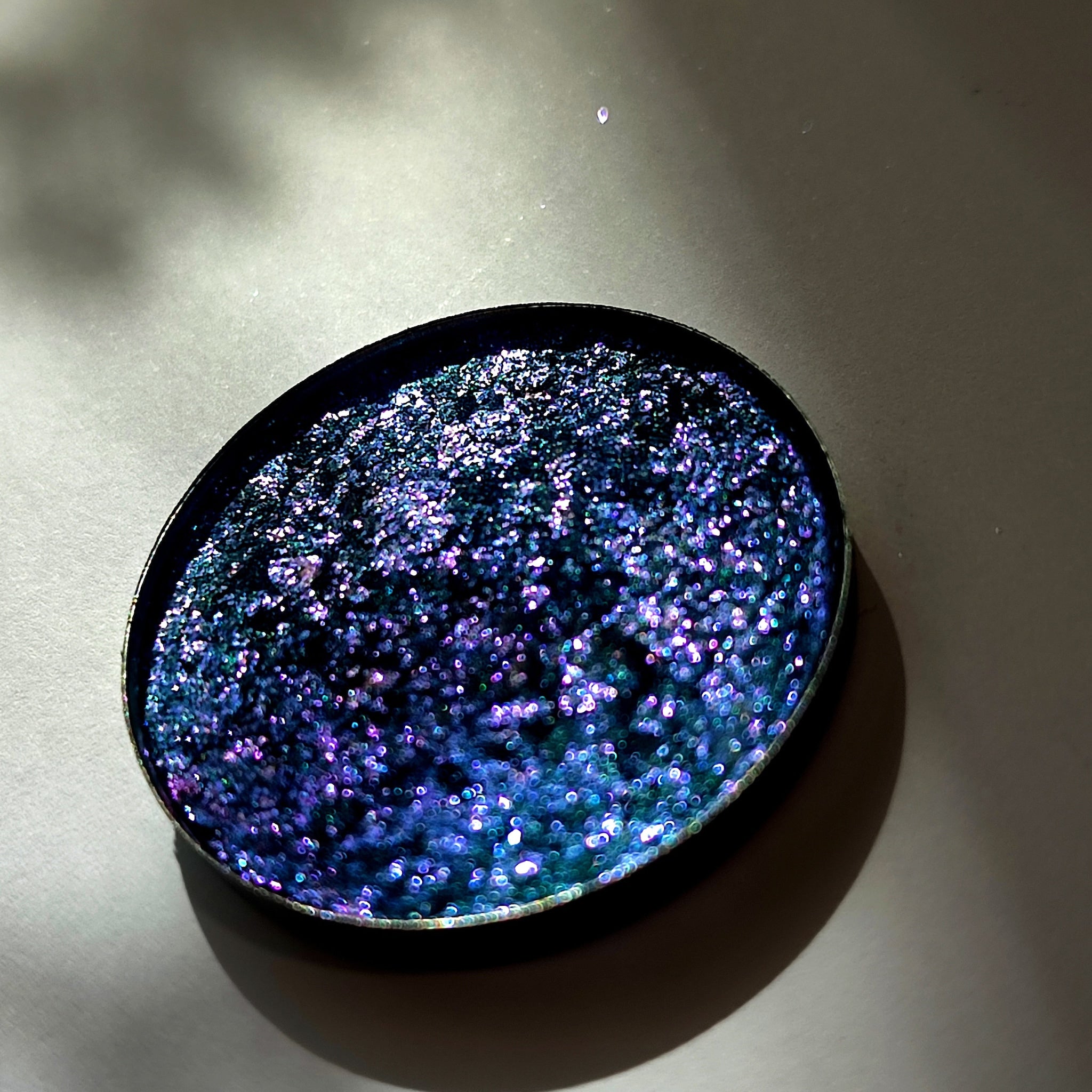 Glitter eyeshadow, oval compact with blue, purple and green iridescent sparkles, black rim