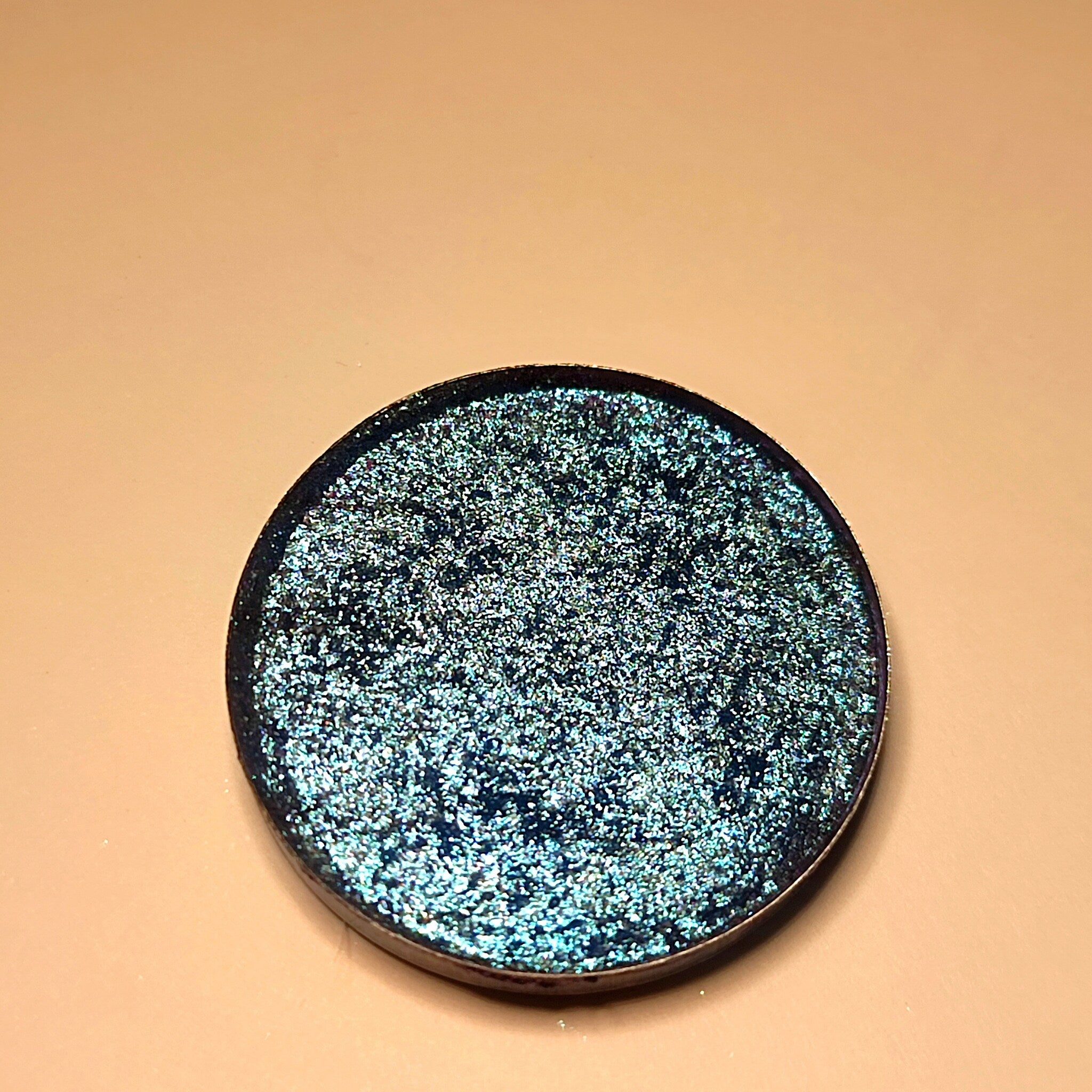 Eyeshadow compact with iridescent green and blue shimmer, dark rim, round shape