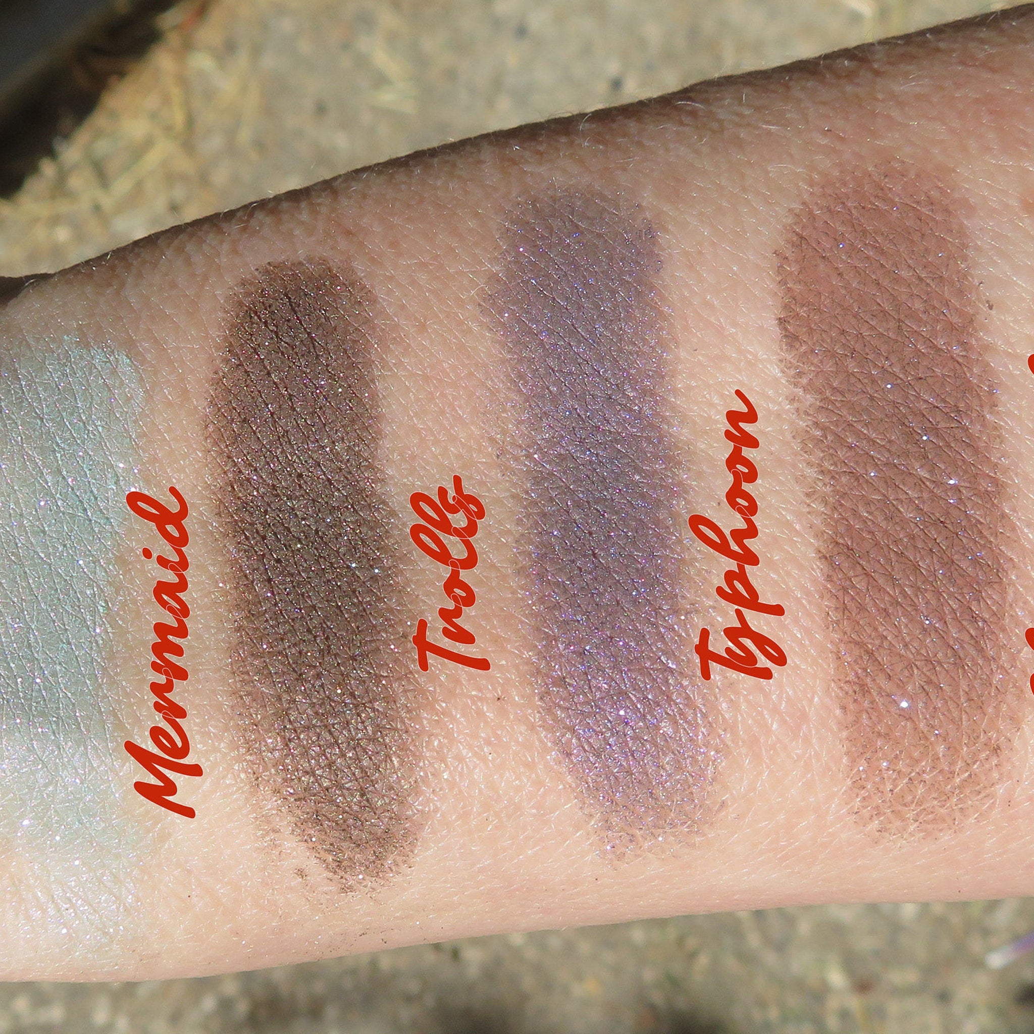 Eyeshadow swatches, Mermaid, brown, purple, and taupe with glitter on arm, names visible