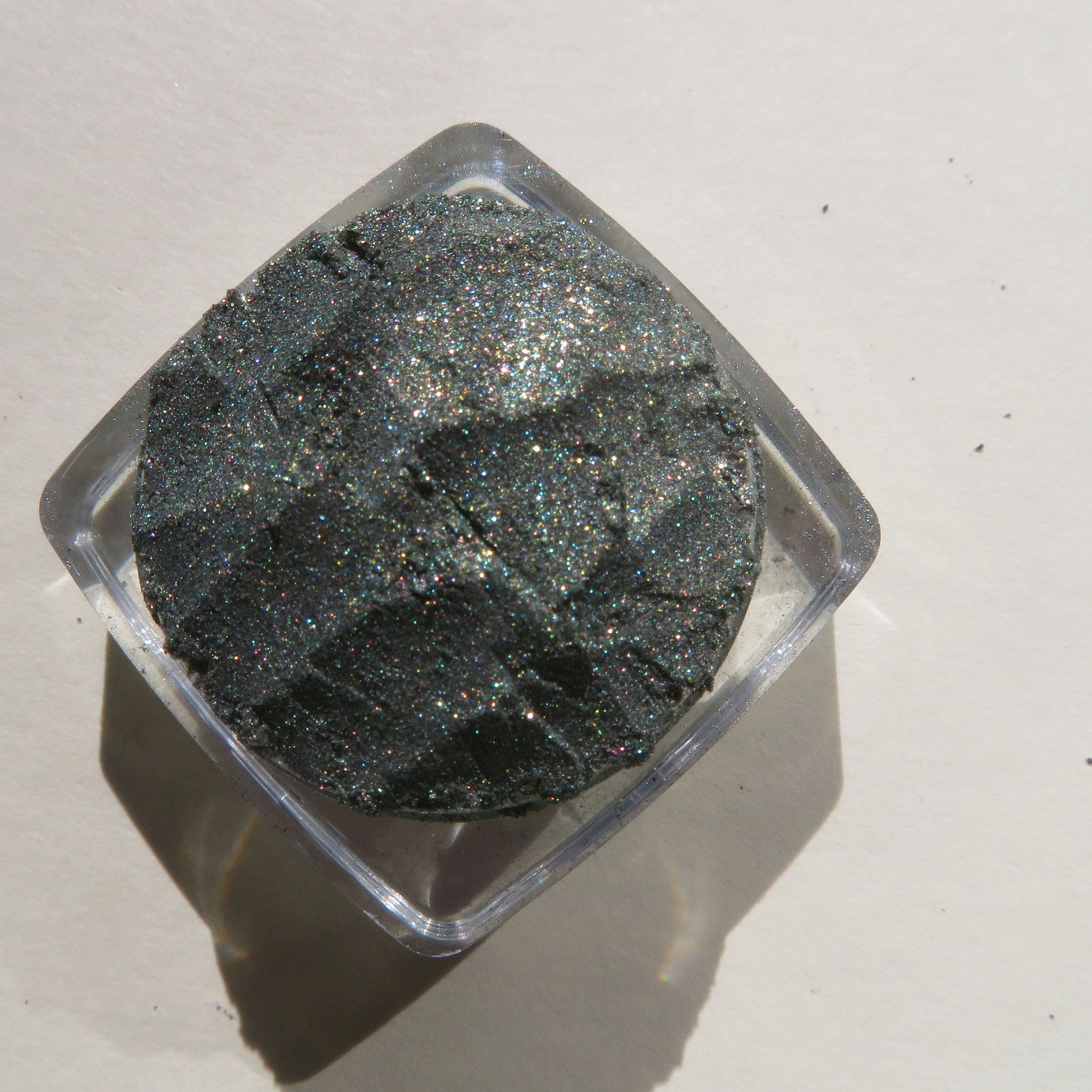 Glitter eyeshadow, dark gray with iridescent shimmer, in a clear square container
