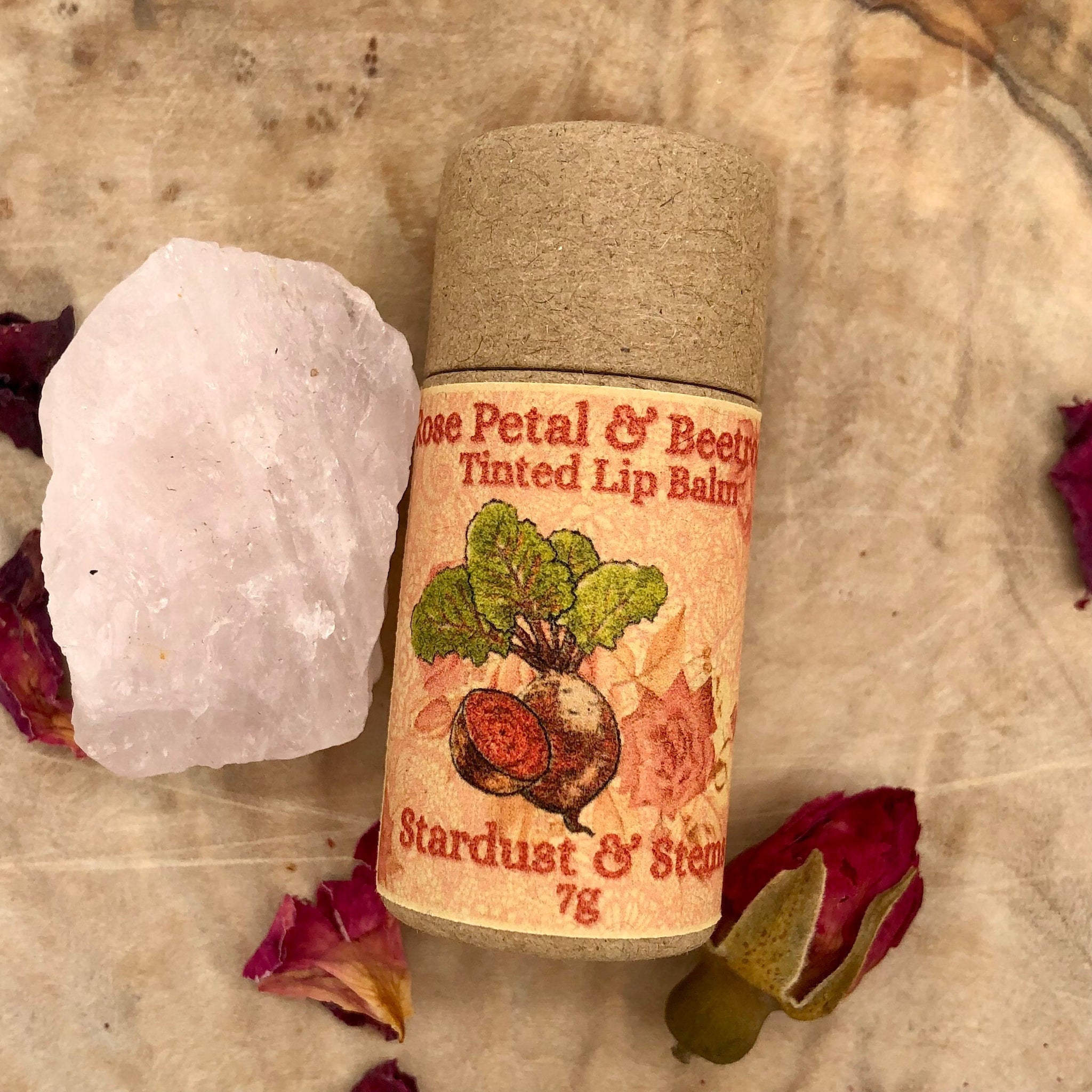 Lip balm tube with beige paper, floral label, and brown cap, next to a pink crystal and rose petals