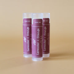 Lip balm tubes with purple label, white text, and clear plastic cap, Lavender + Orange flavor