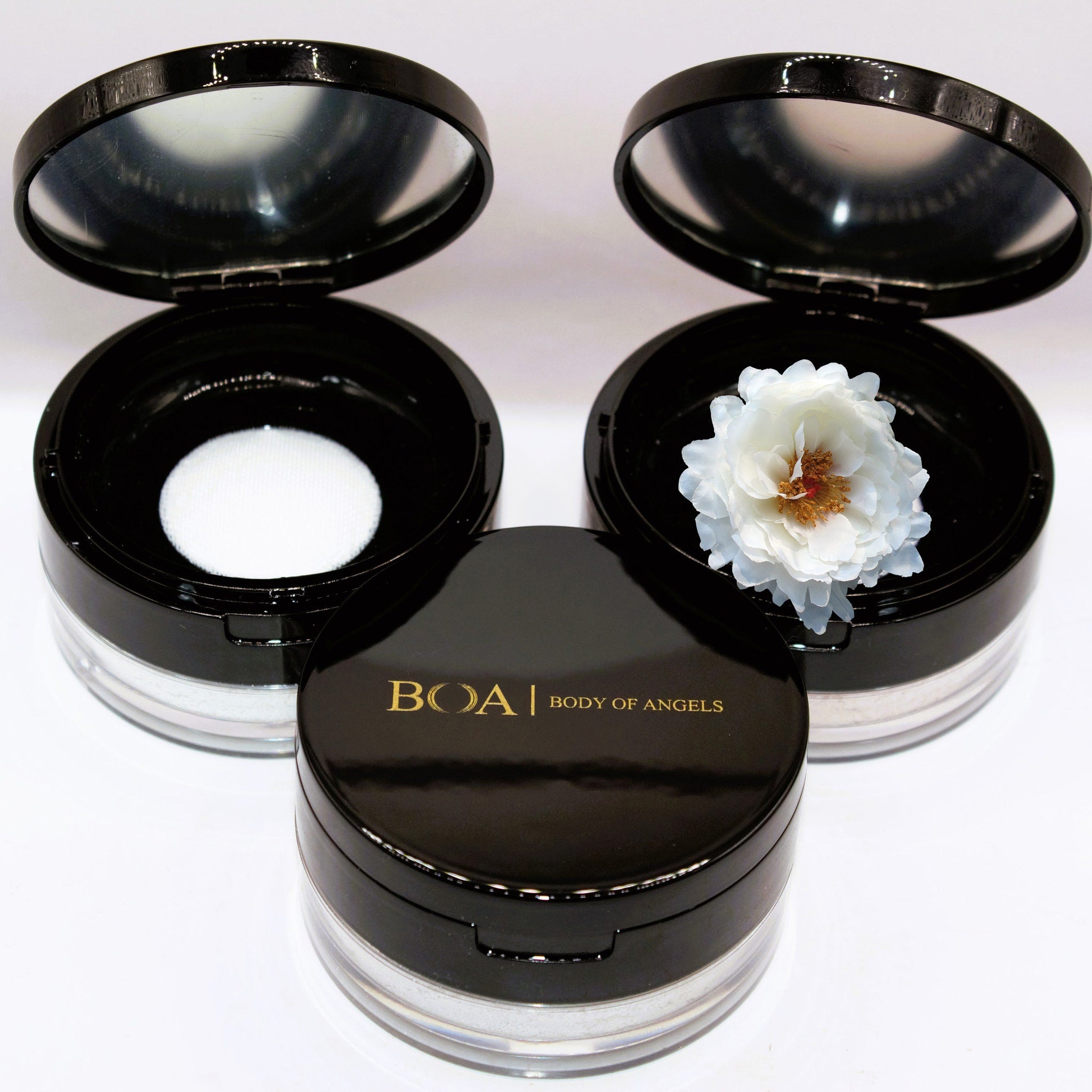 Face powder compacts, black glossy lids, clear base, white powder, and a flower inside