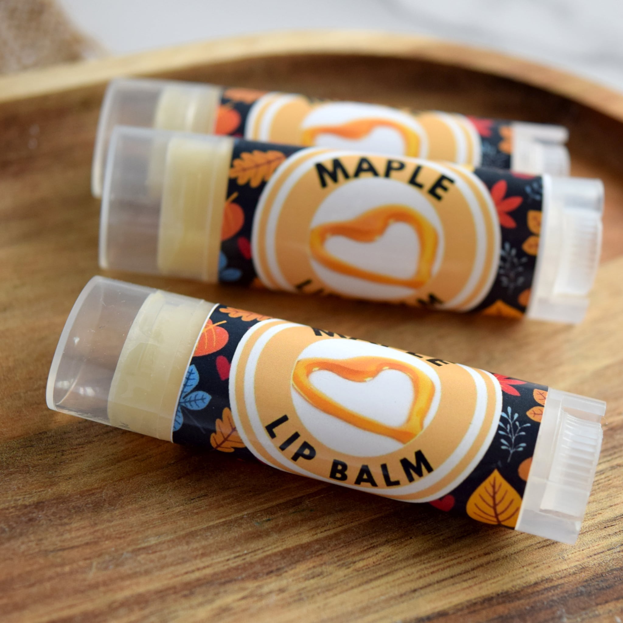Lip balm tubes with maple label, clear plastic, and autumn leaf pattern on wood surface
