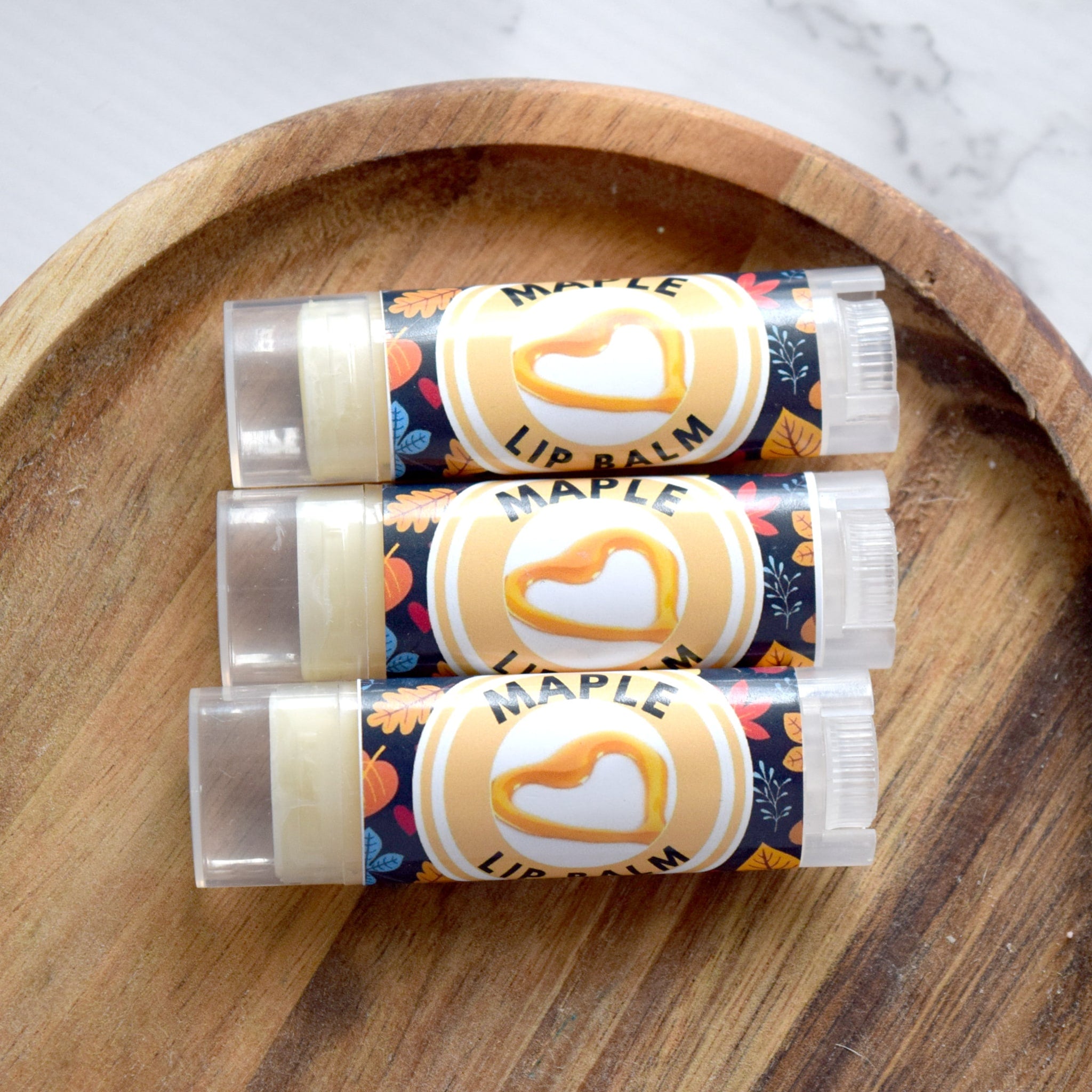Lip balm tubes with "Maple" text, clear plastic, and autumn leaf pattern on wooden tray