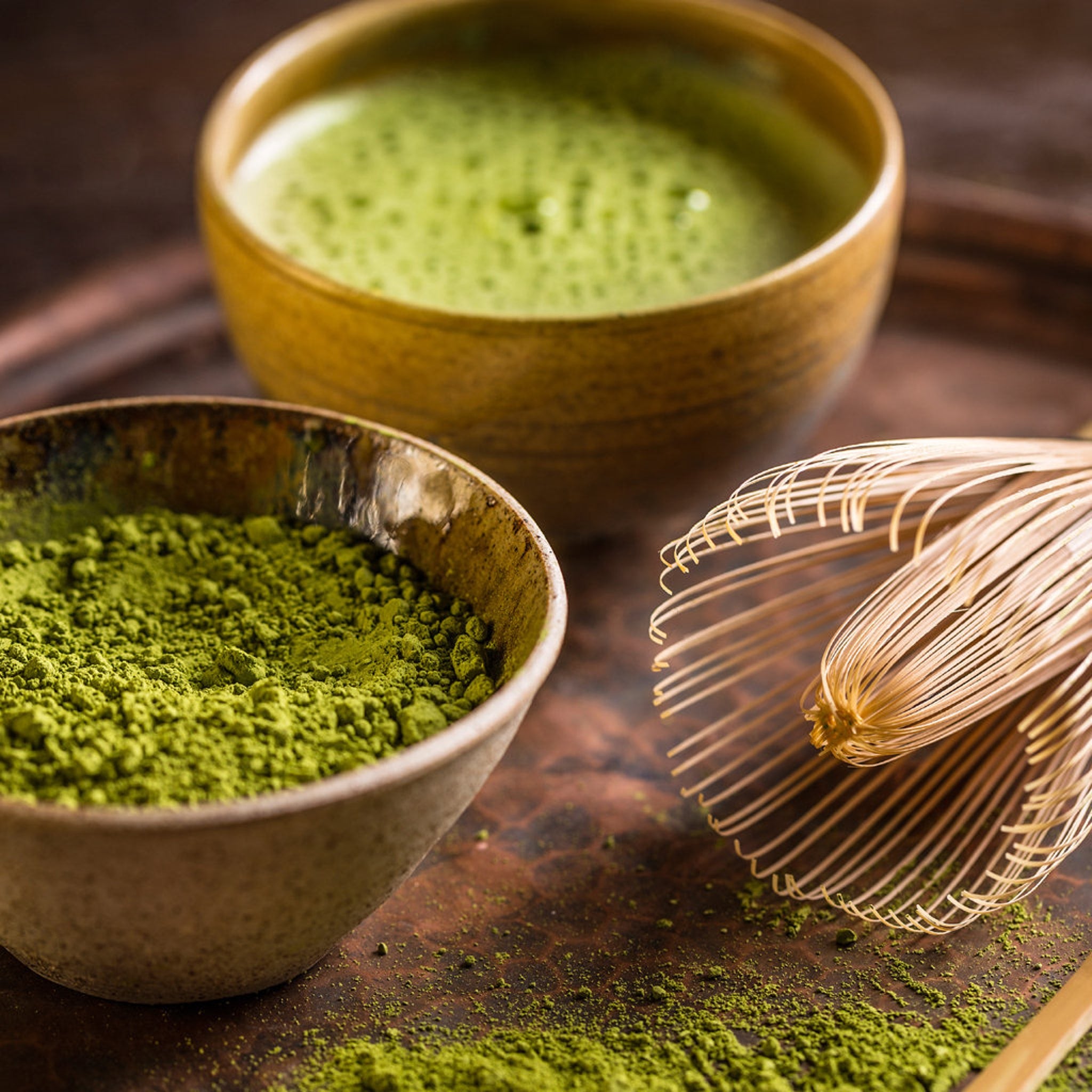Matcha tea with green powder in a brown bowl and whisk, on a wooden surface