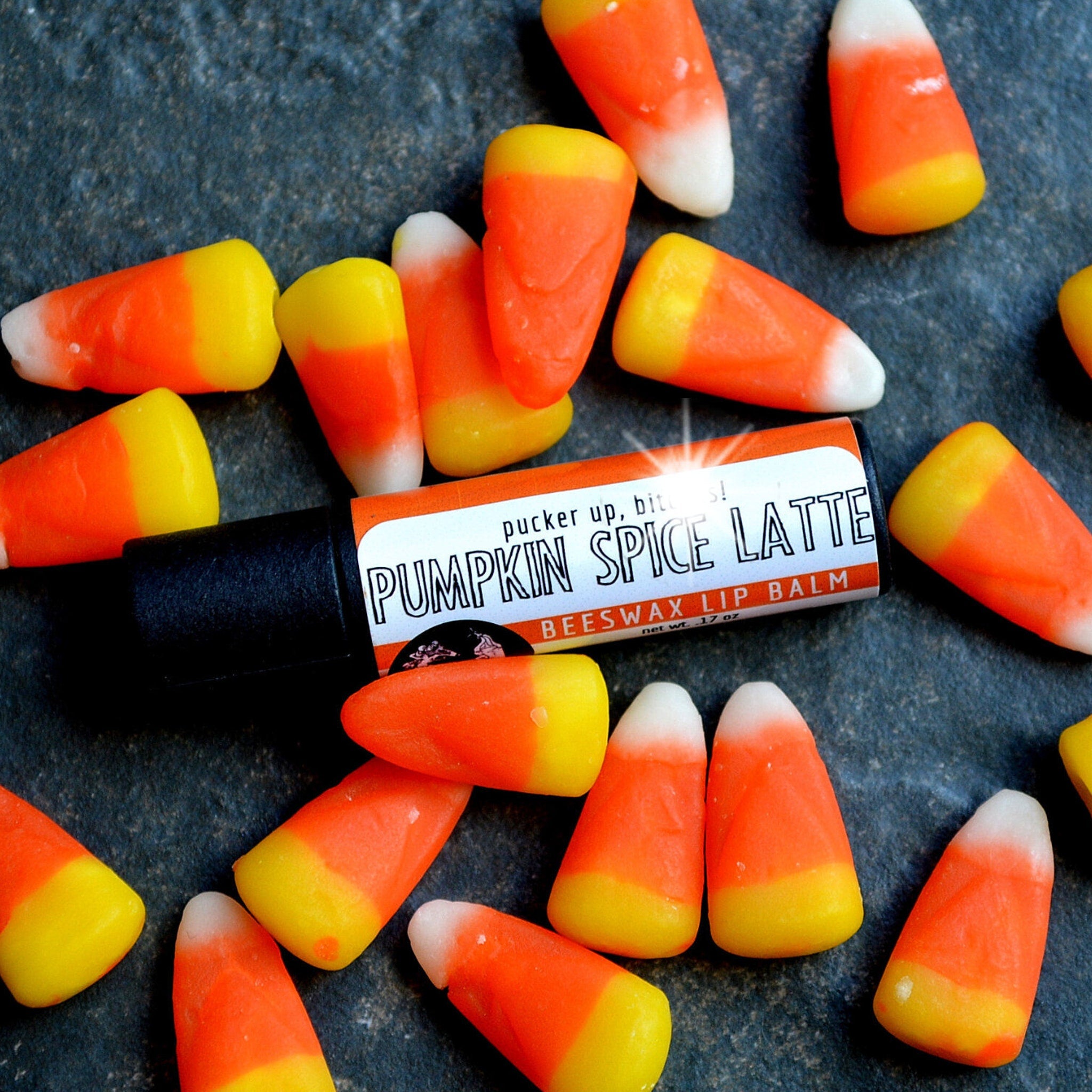 Lip balm tube with black base, orange and white label, surrounded by candy corn on textured surface