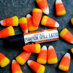 Lip balm tube with black base, orange and white label, surrounded by candy corn on textured surface