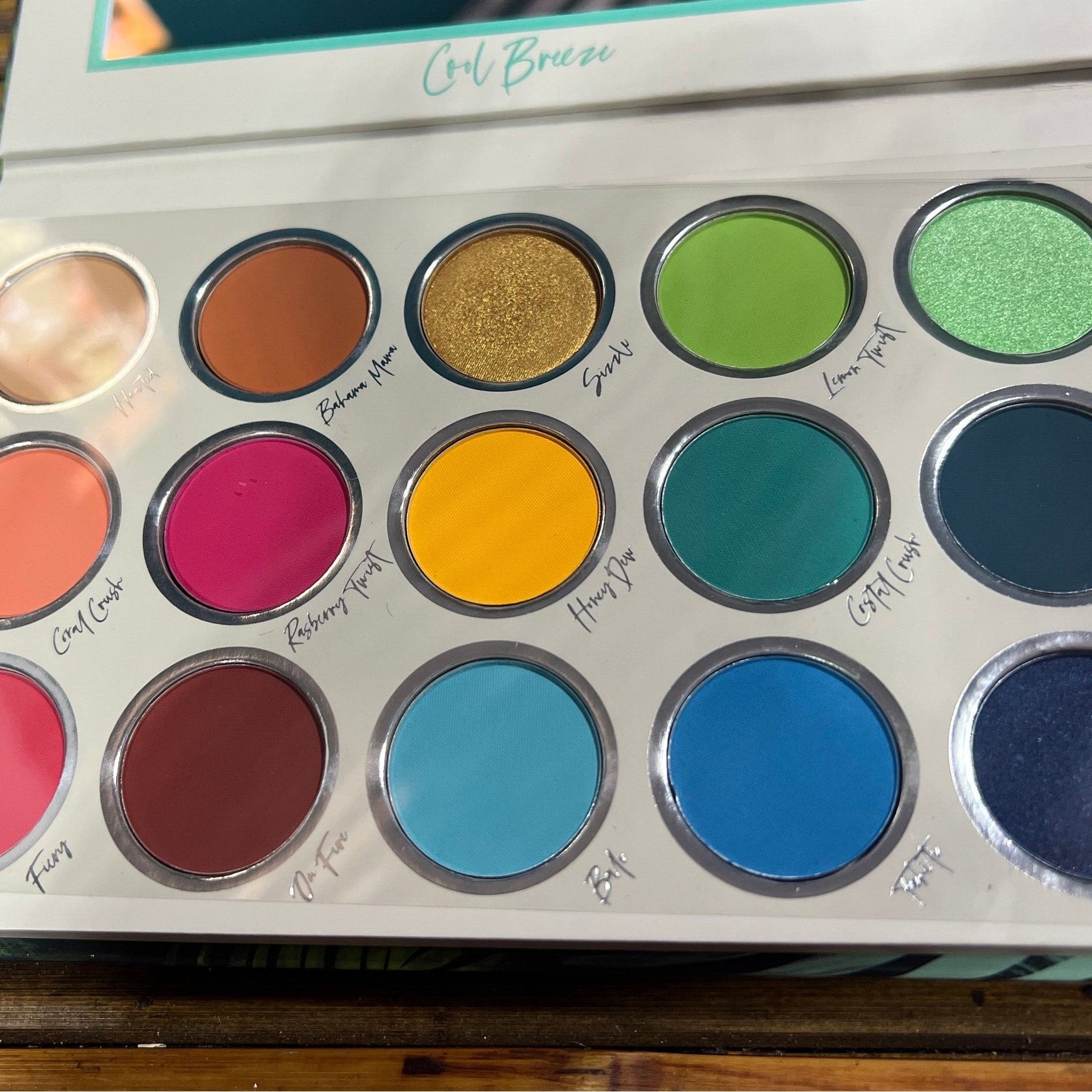 Eyeshadow palette with various colors, including green, blue, and gold, in a white case labeled Cool Breeze