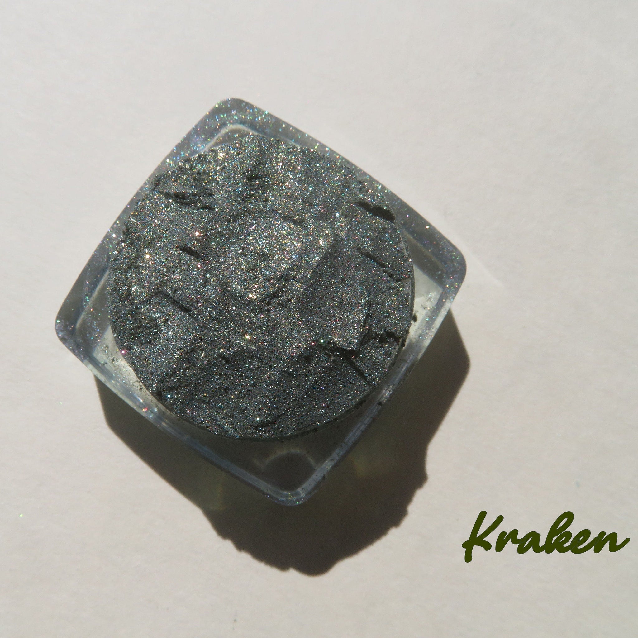 Eyeshadow pot with gray glitter, clear plastic container, and Kraken text on white background