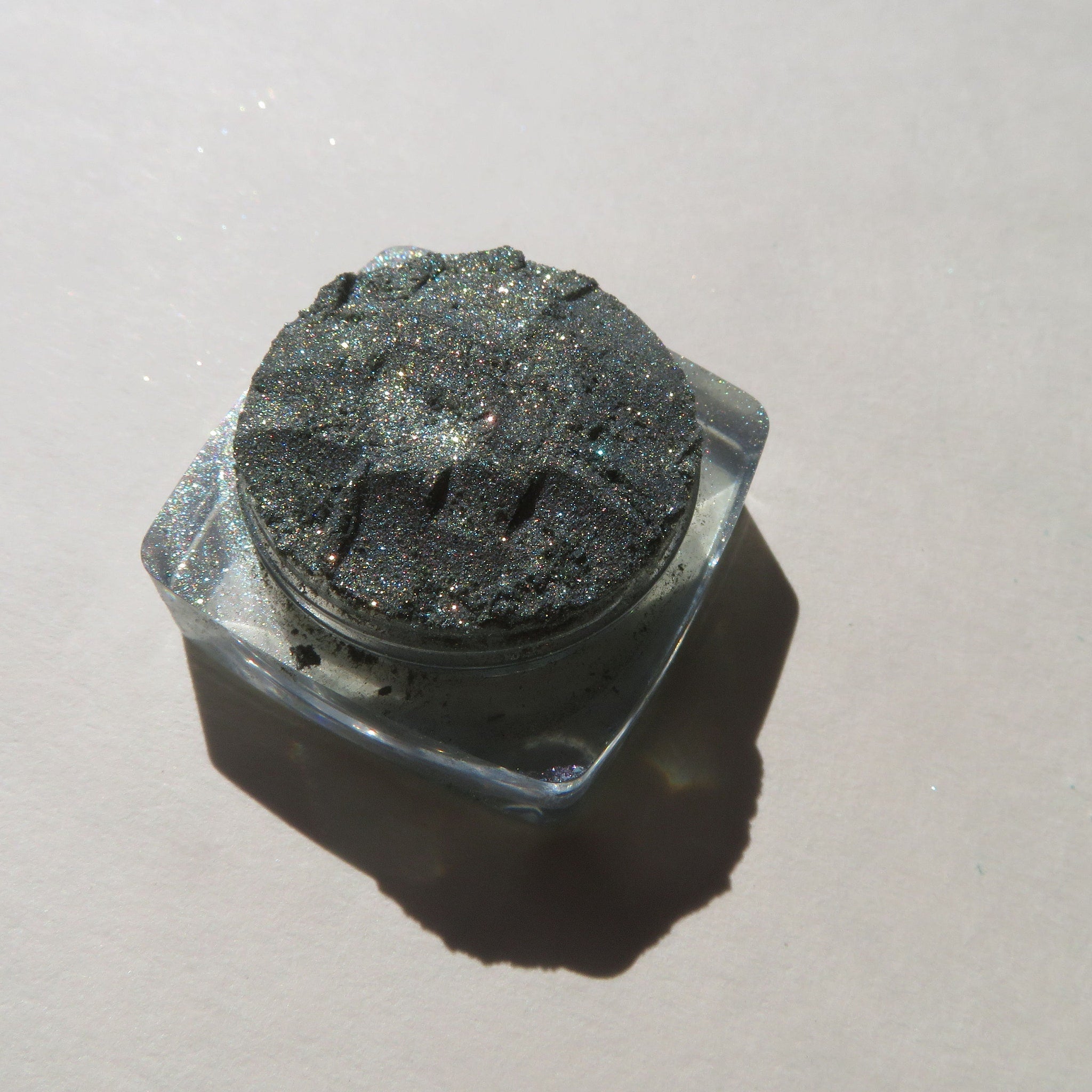 Glitter eyeshadow in dark gray with sparkling particles in a clear plastic container, top view