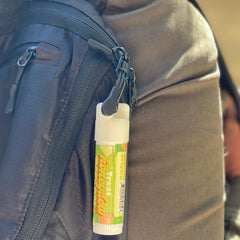 Lip balm tube with white, green and orange label, attached to black backpack with clasp