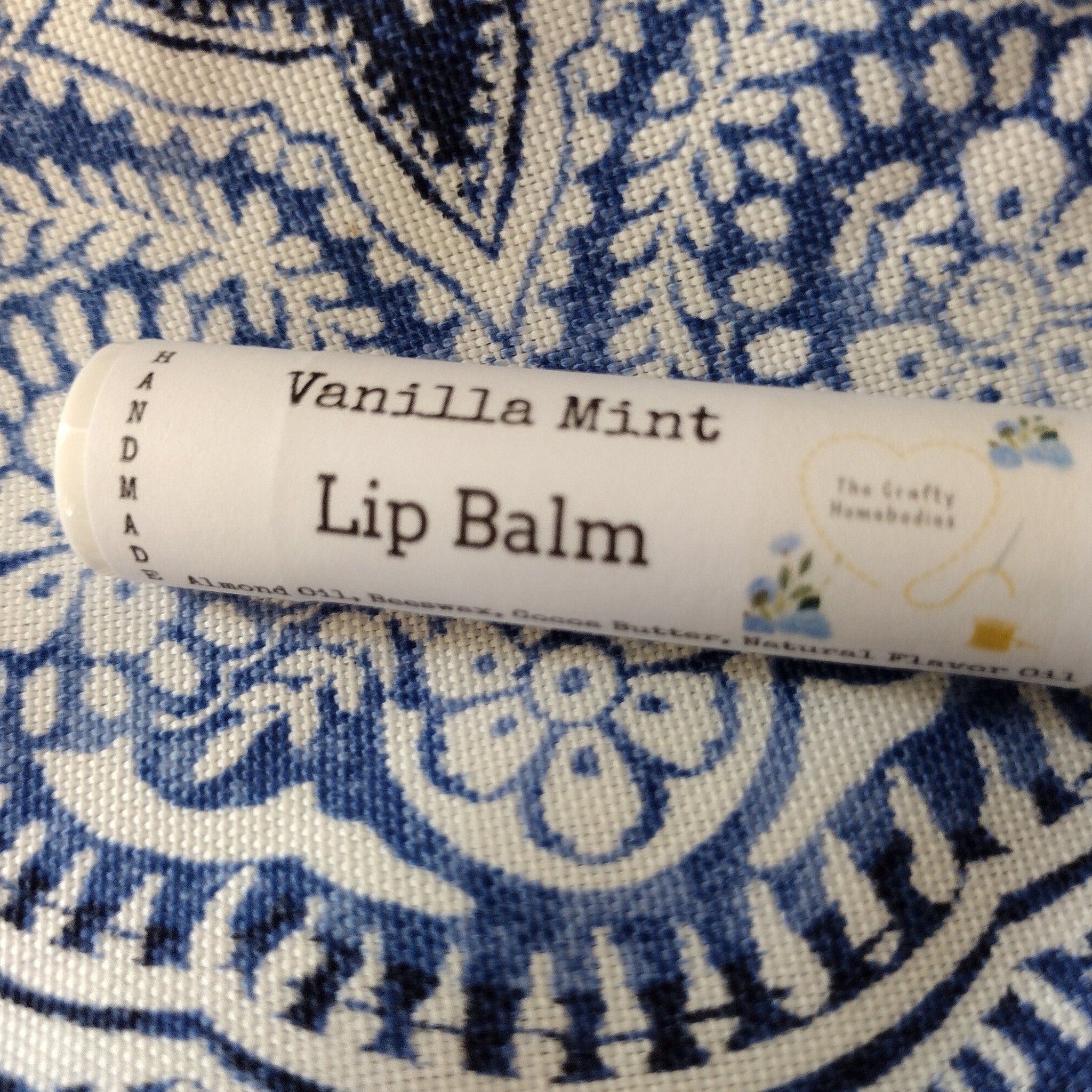 Lip balm tube with white label, black text, and blue floral pattern on fabric background