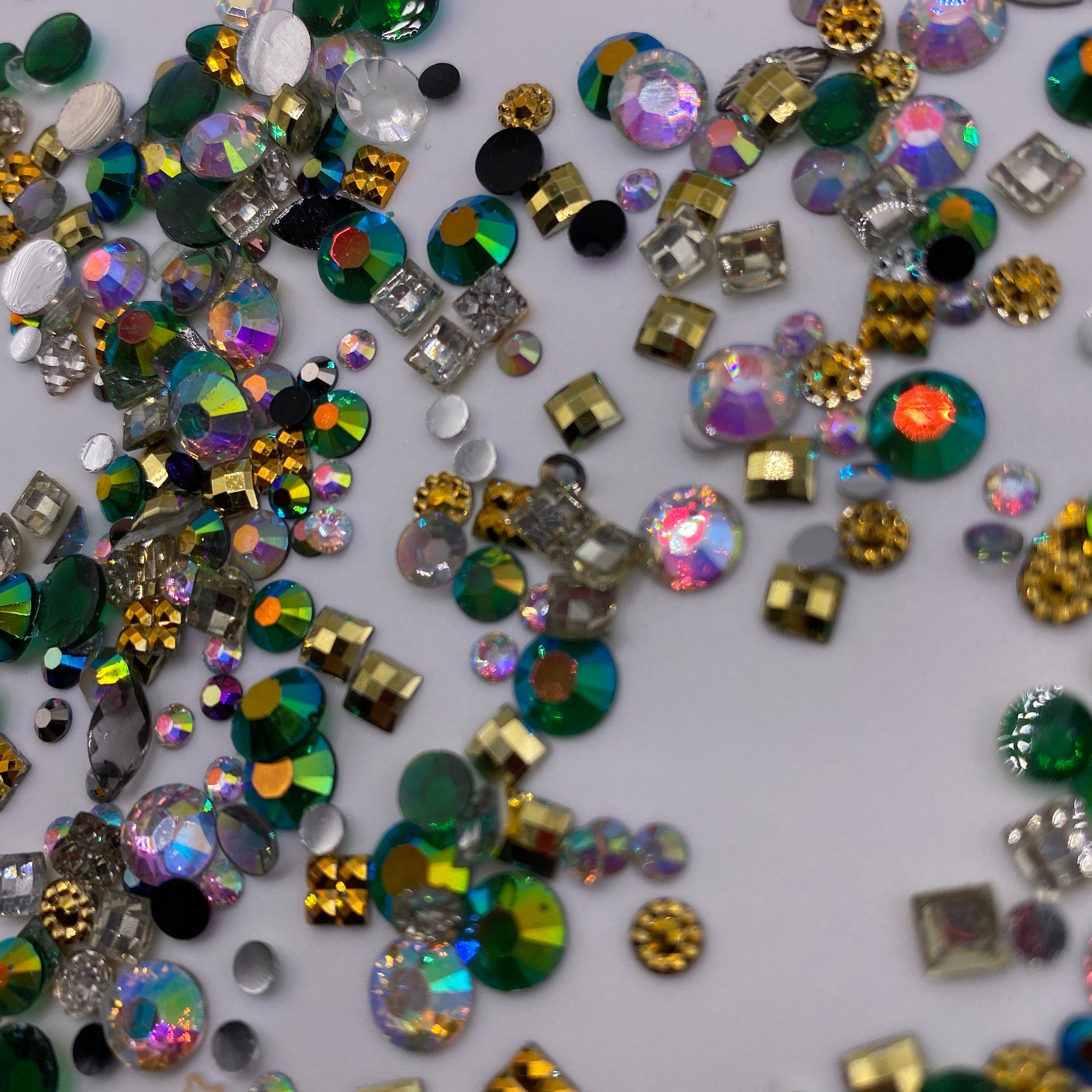 Rhinestones in various shapes, iridescent, green, and gold colors scattered on white surface