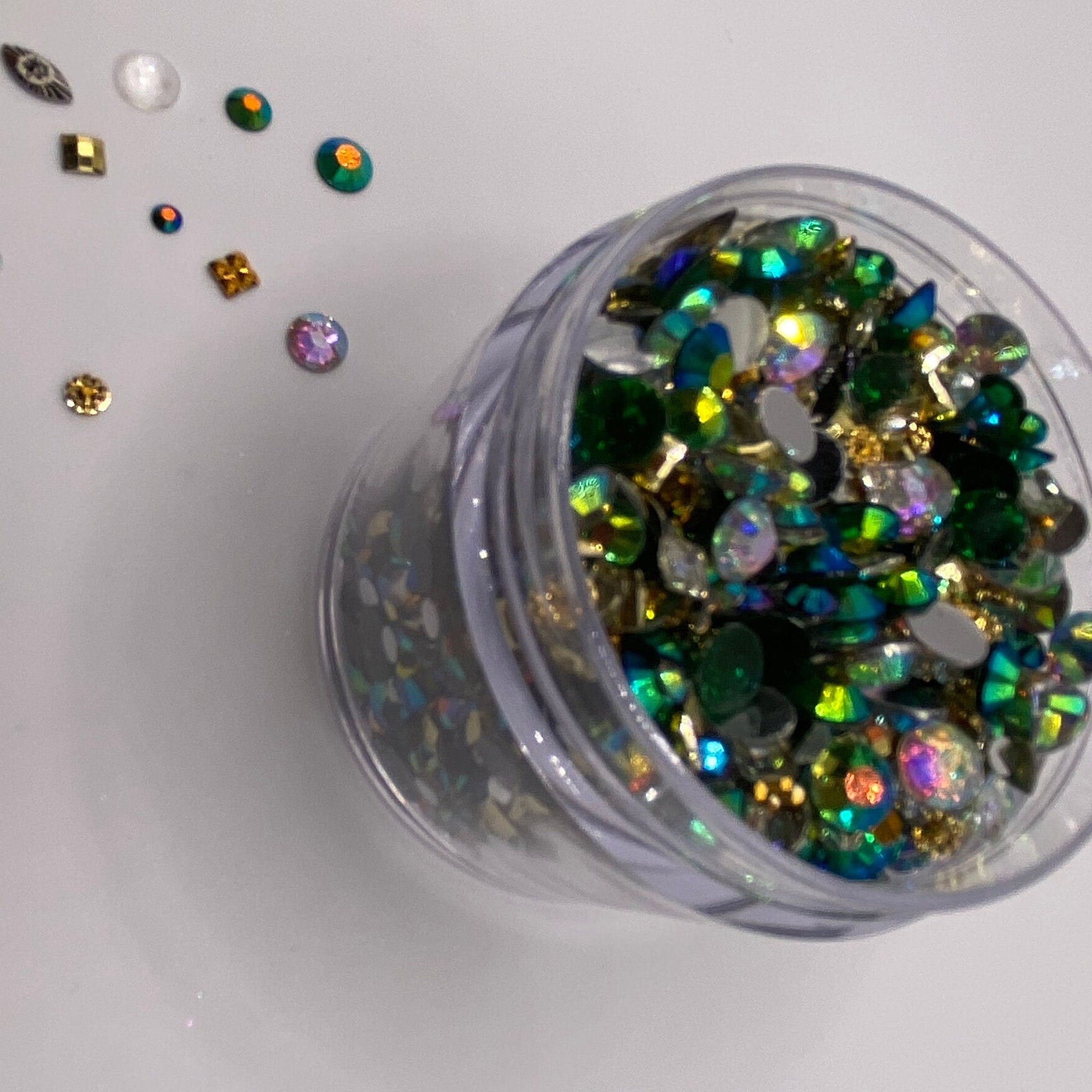 Rhinestones in a clear container, with green, gold, and iridescent colors, scattered on white surface