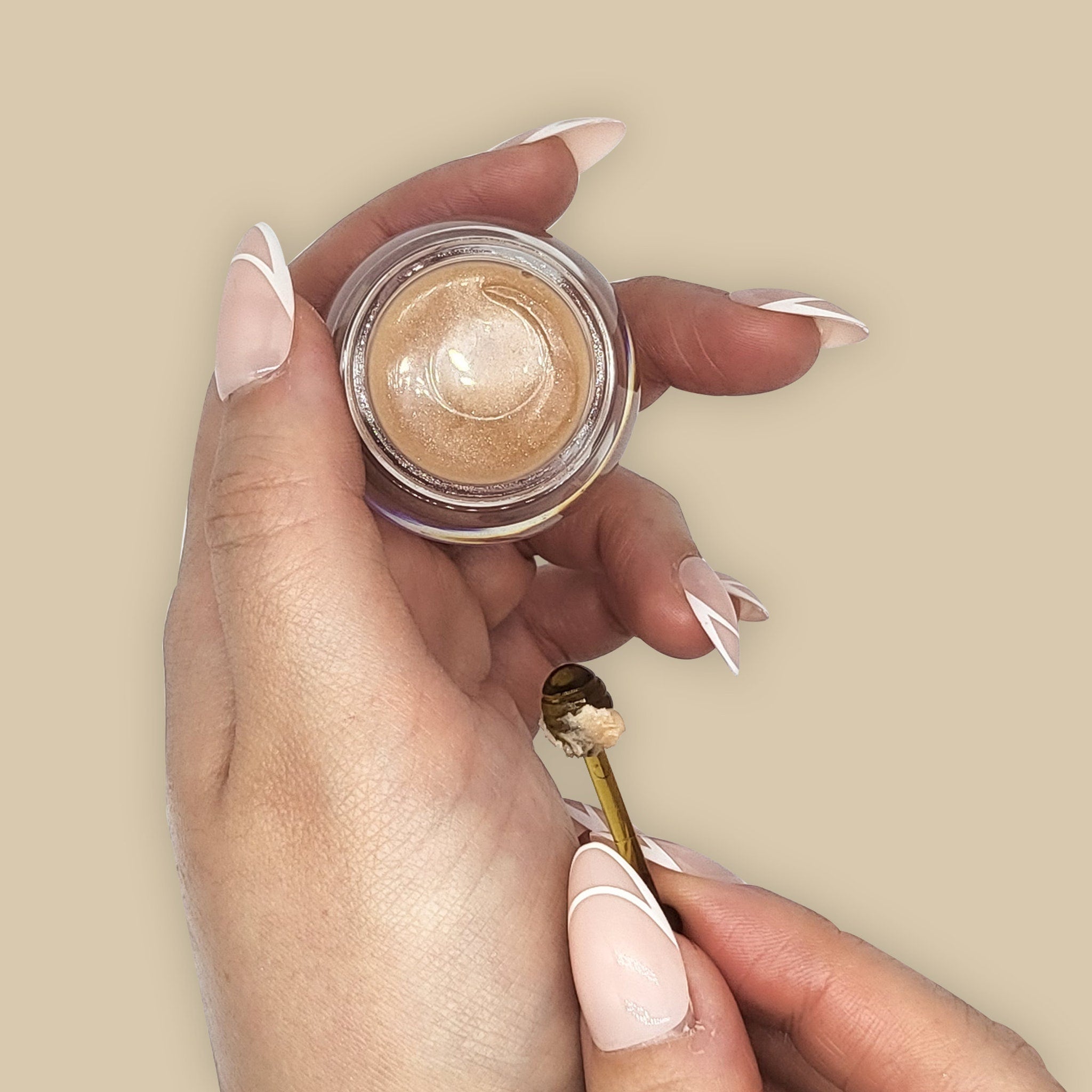 Gold eyeshadow in a clear jar held by hands with nude and white nail art, and a gold tool