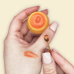 Orange lip balm in a yellow jar and on a hand, with a small applicator spoon