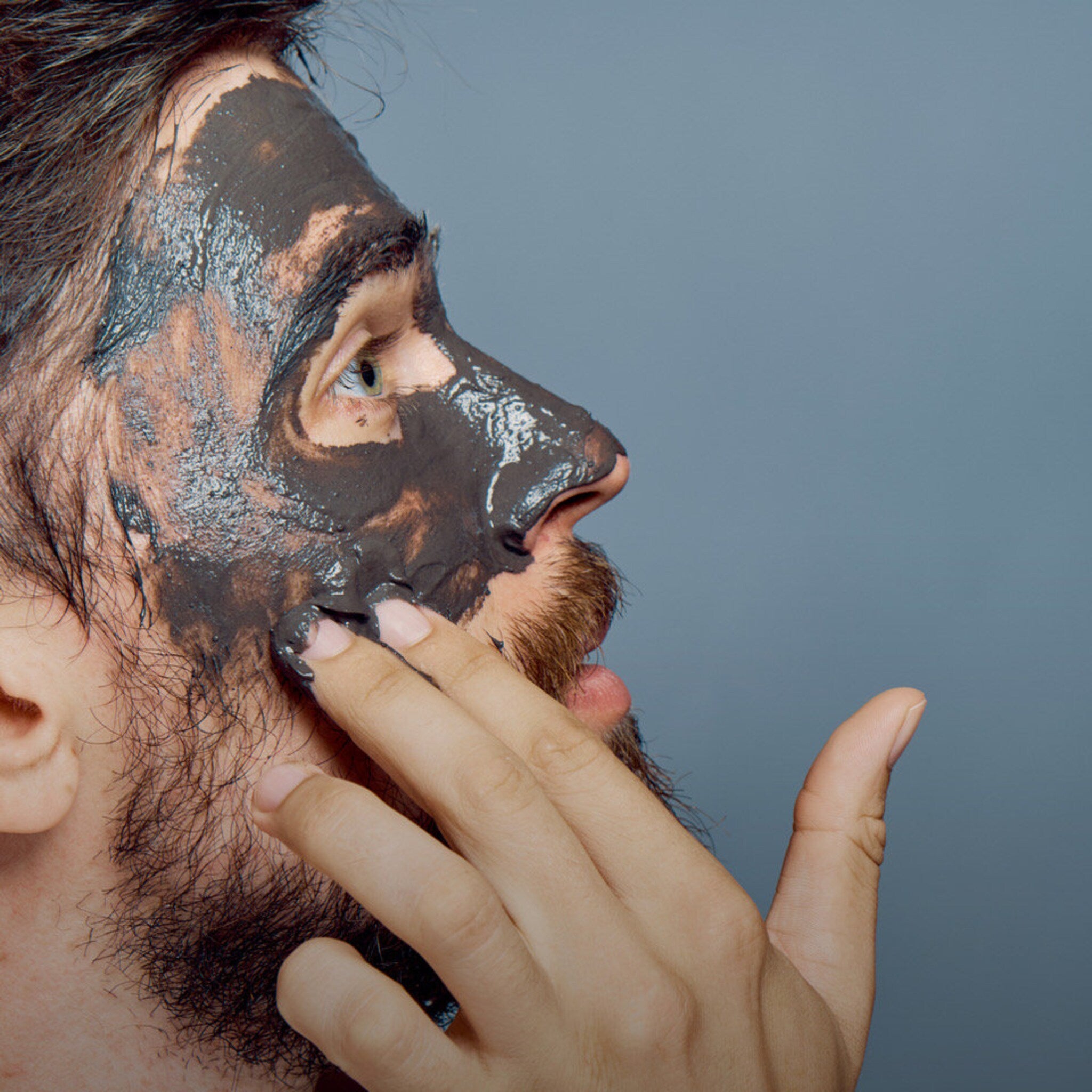 Face mask application, dark gray mud mask, applied to face, with hand touching skin