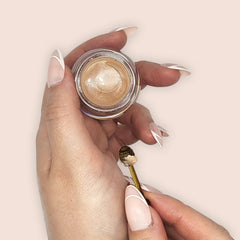 Gold eyeshadow in a glass jar, held by hands with pink and white nails, with a small applicator