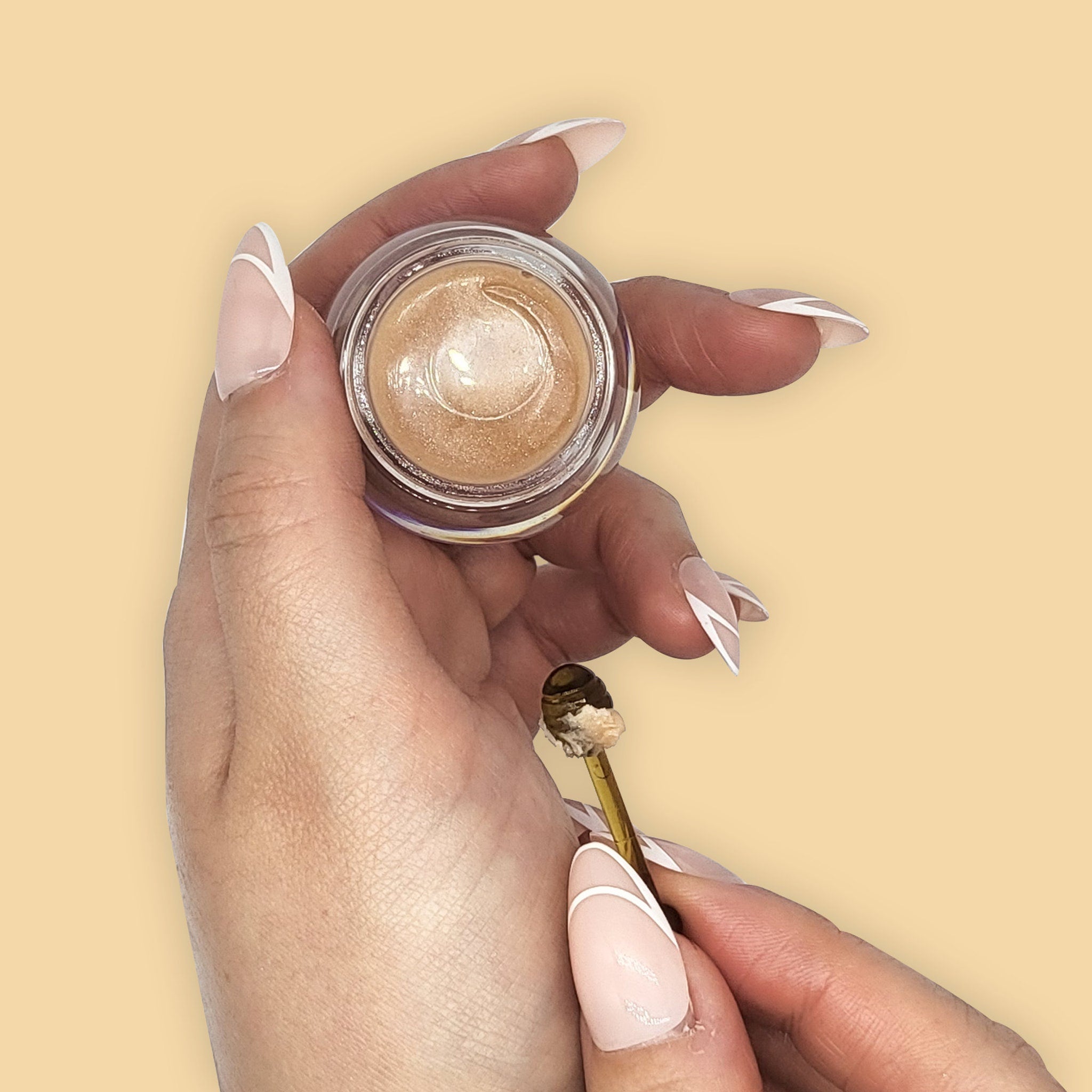 Cosmetic product in clear jar with gold contents, held by hands with light pink nails and white tips