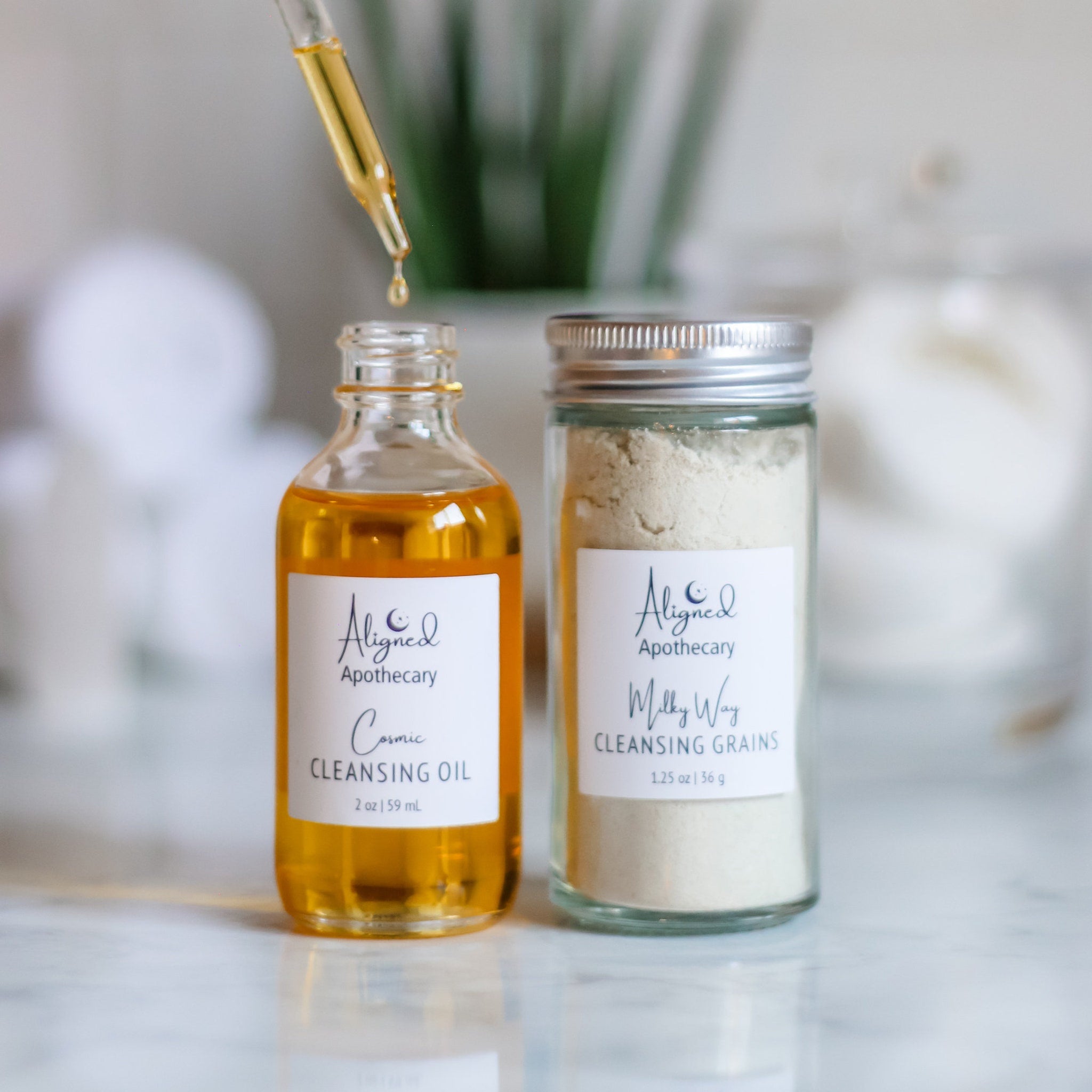 Cleansing oil and grains with glass bottles, yellow liquid, and white labels with Aligned Apothecary text