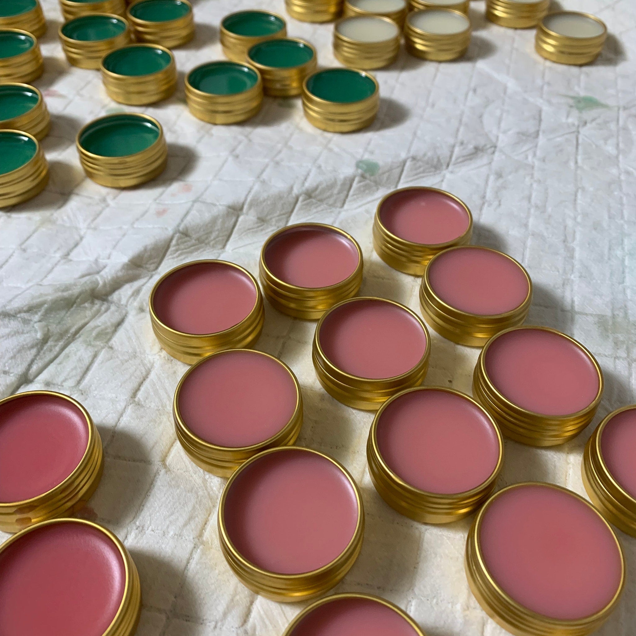 Lip balm tins, gold metal containers with pink and green balm, arranged on white fabric