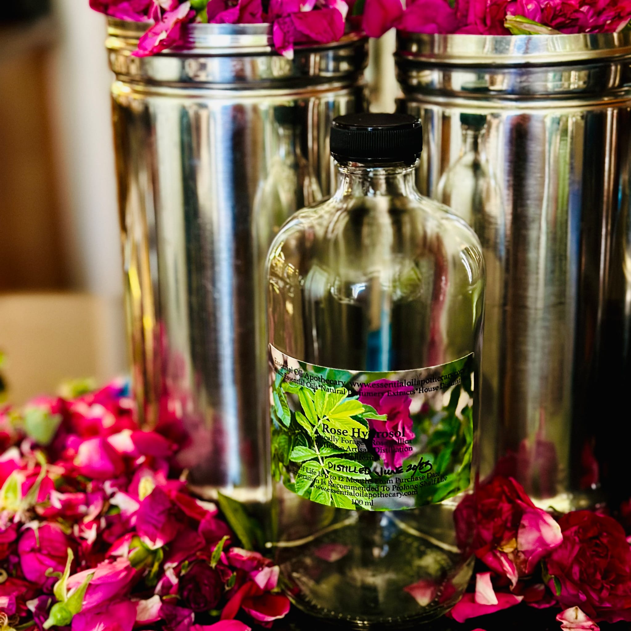 Rose hydrosol bottle with clear glass, black cap, and floral label, surrounded by pink rose petals