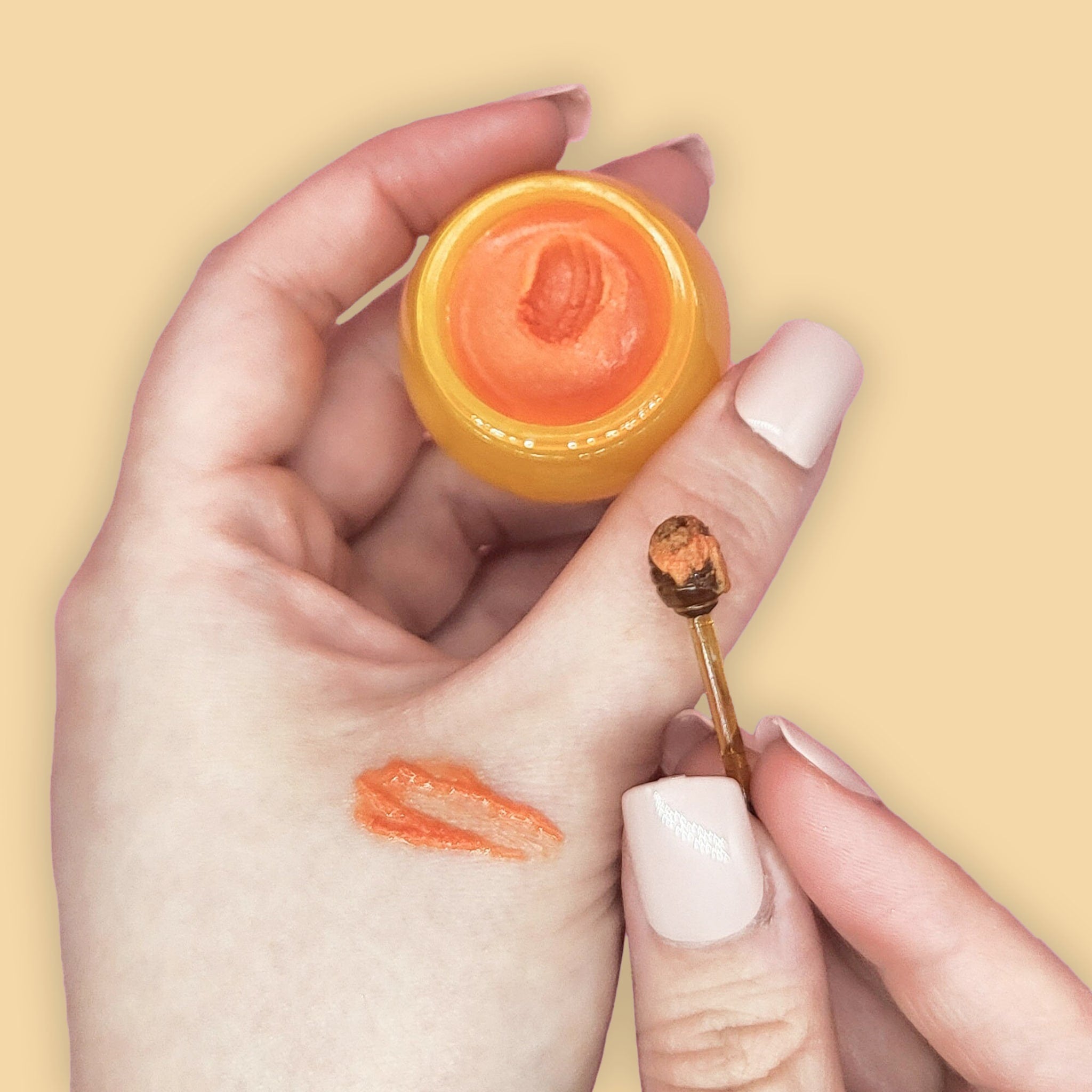 Lip balm orange color in a jar and applied on skin, with applicator tool