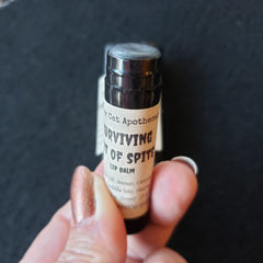 Lip balm tube with black and white label, held by hand, text "Surviving out of Spite" visible