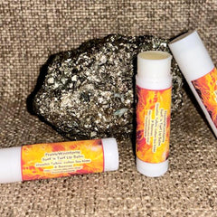 Lip balms with white tubes, orange and yellow labels, and text, near a rock on fabric