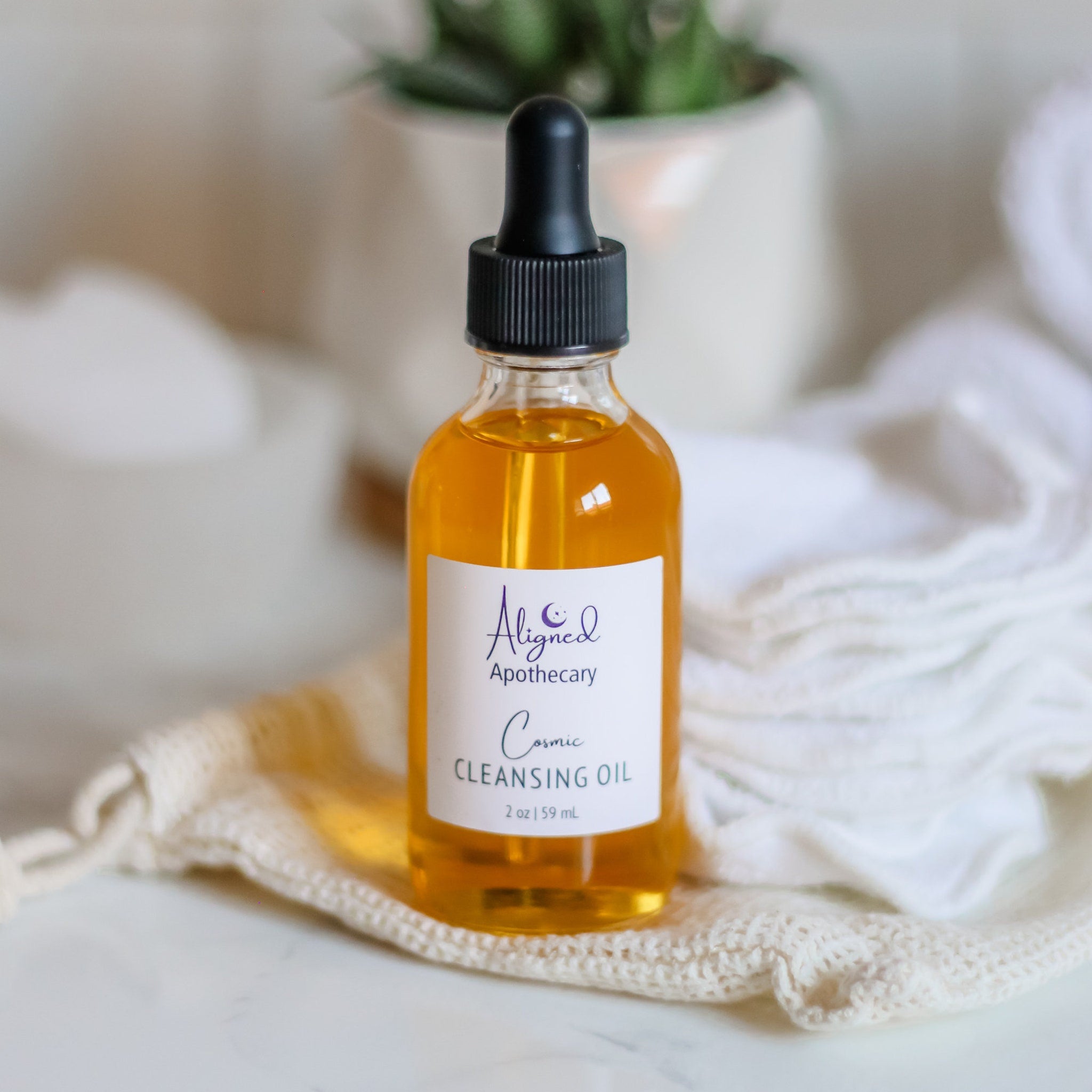 Cleansing oil bottle with amber liquid, black dropper, and Aligned Apothecary label on white fabric