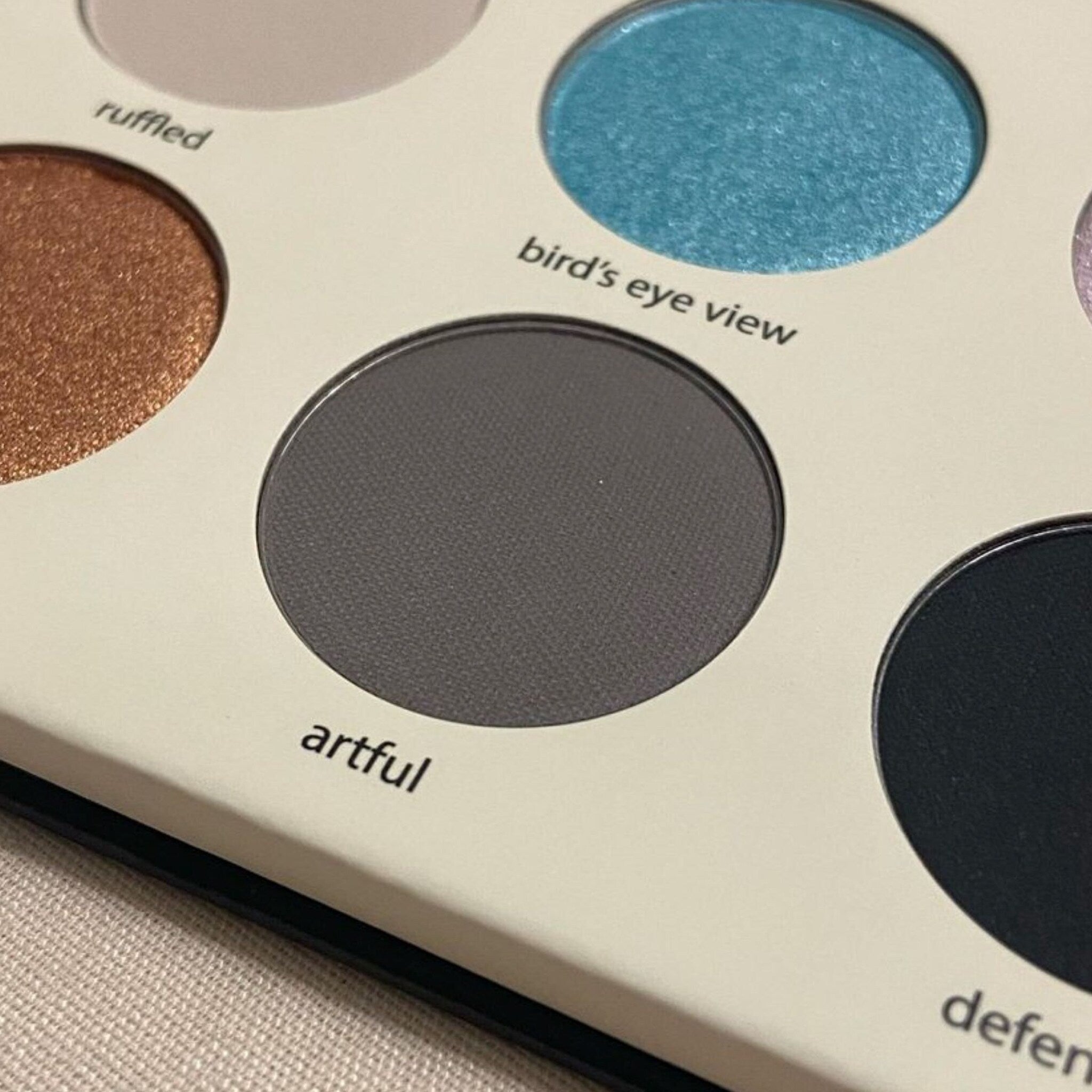 Eyeshadow palette with gray, blue, and black shades in round pans, with white background and text labels.