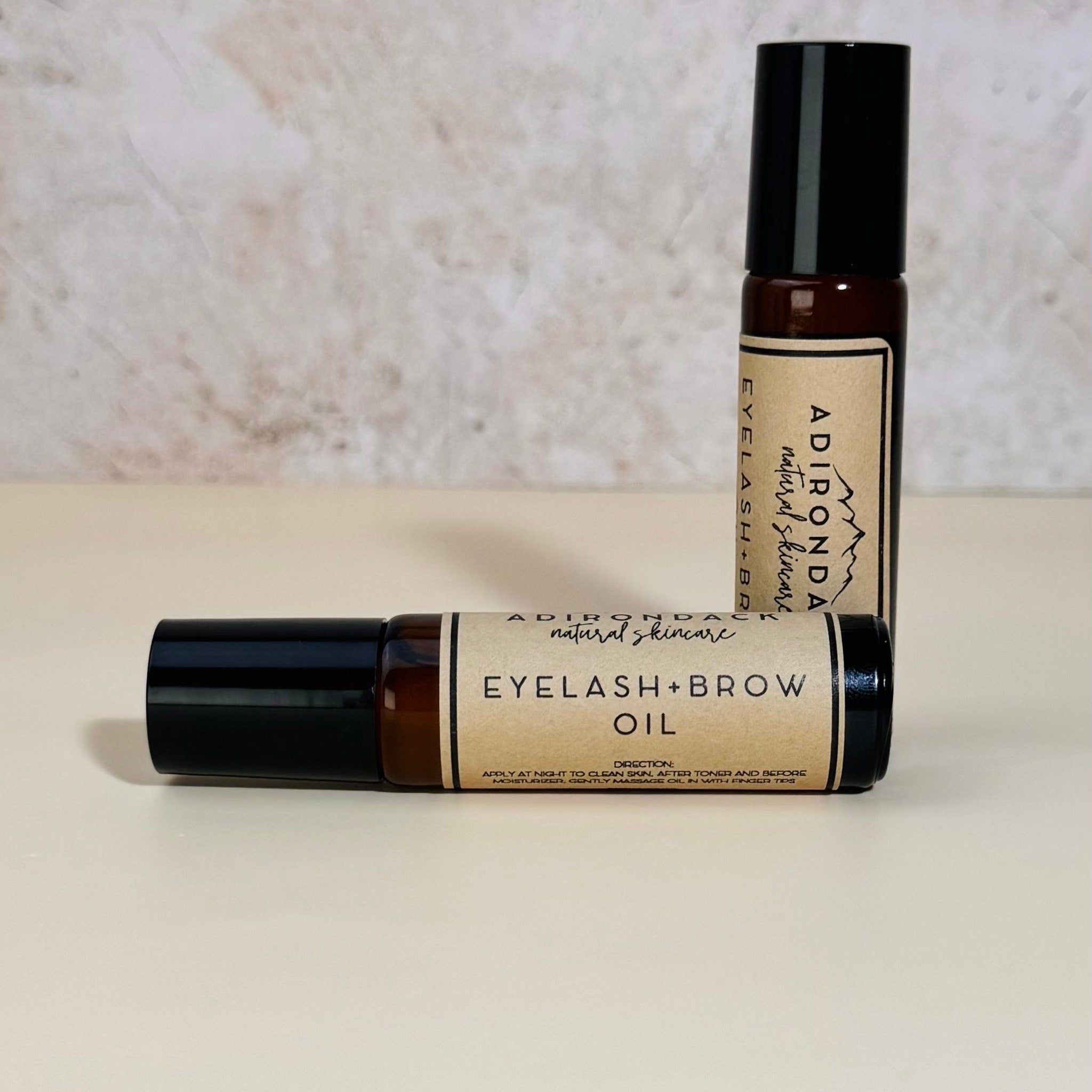 Eyelash and brow oil in brown glass bottles with black caps, labeled Adirondack natural skincare, on a white surface