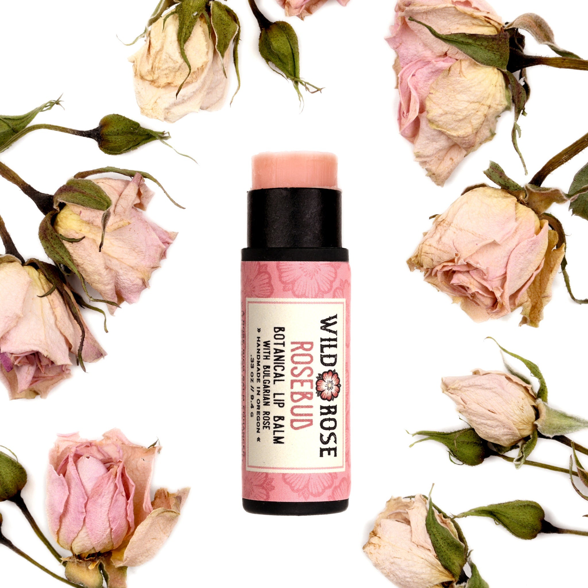 Lip balm tube with pink label, black cap, surrounded by dried pink roses and buds, Wild Rose brand