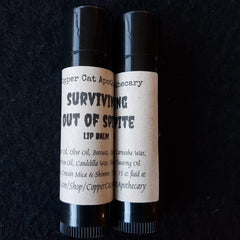 Lip balm tubes with black caps and paper labels, "Surviving Out Of Spite" brand name visible