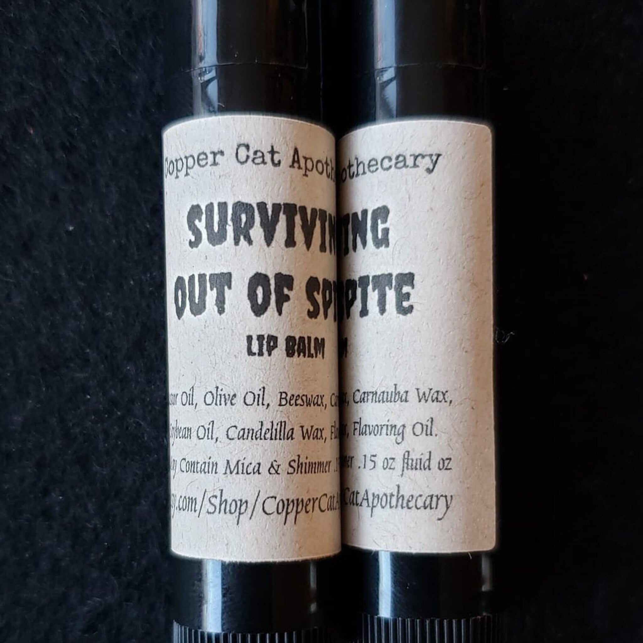 Lip balm tubes with black base and paper labels, Copper Cat Apothecary text visible, "Surviving Out of Spite" text