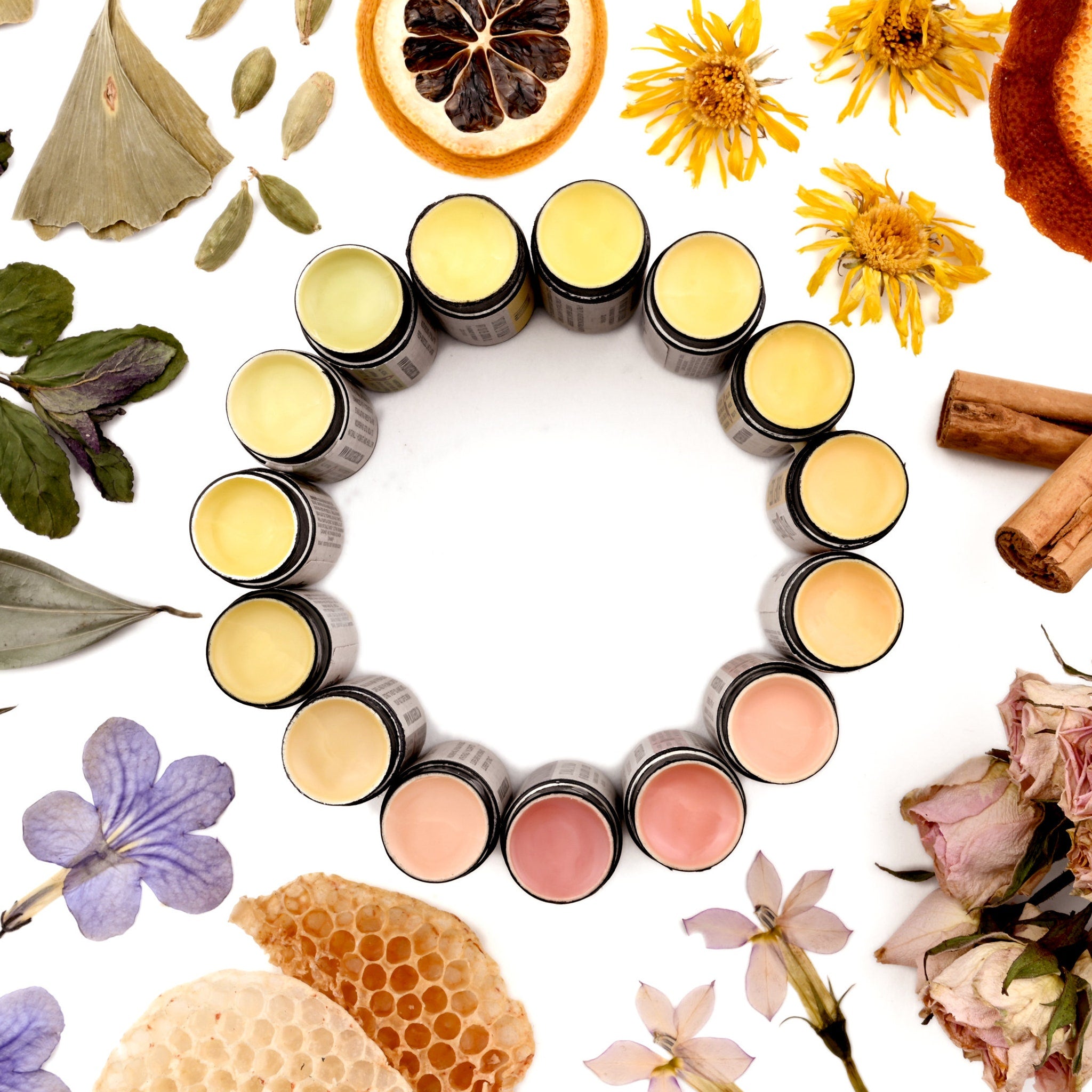 Lip balms in small black jars with various shades of yellow and pink, arranged in a circle with flowers