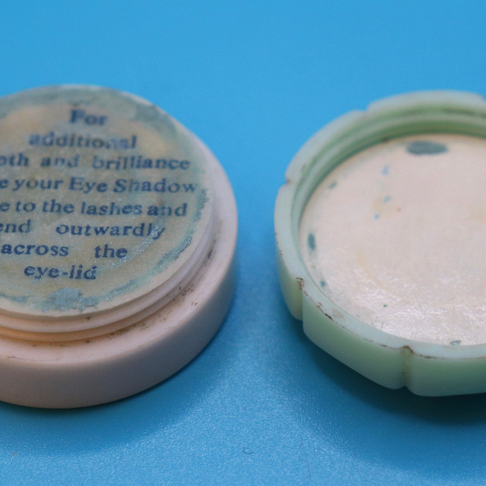 Eye shadow container, off-white and green plastic, open lid, blue text on top, on blue surface