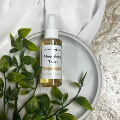 Nourishing toner bottle with white label, clear liquid, and green leaves on a gray plate