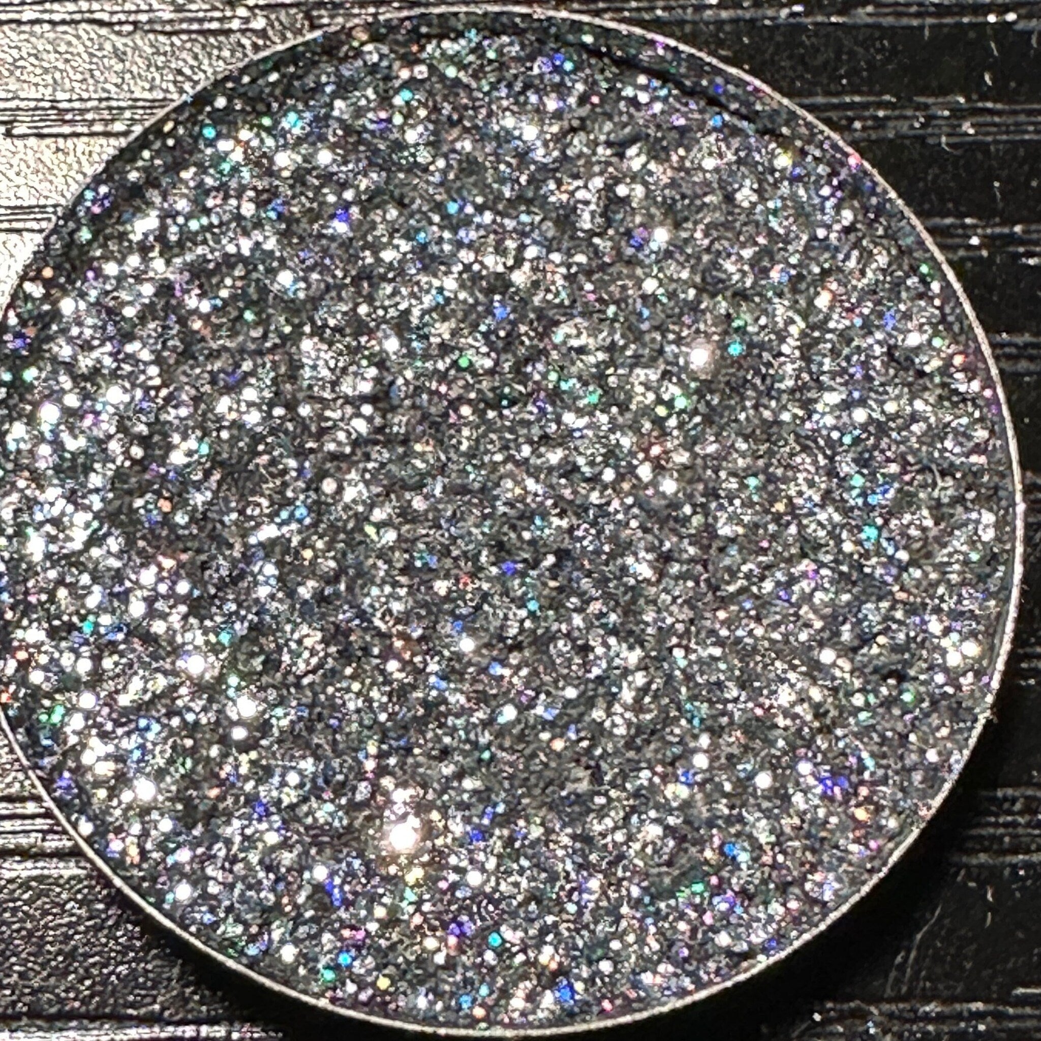Close-up of dark gray holographic glitter eyeshadow in a round 26mm metal pan with reflective sparkles