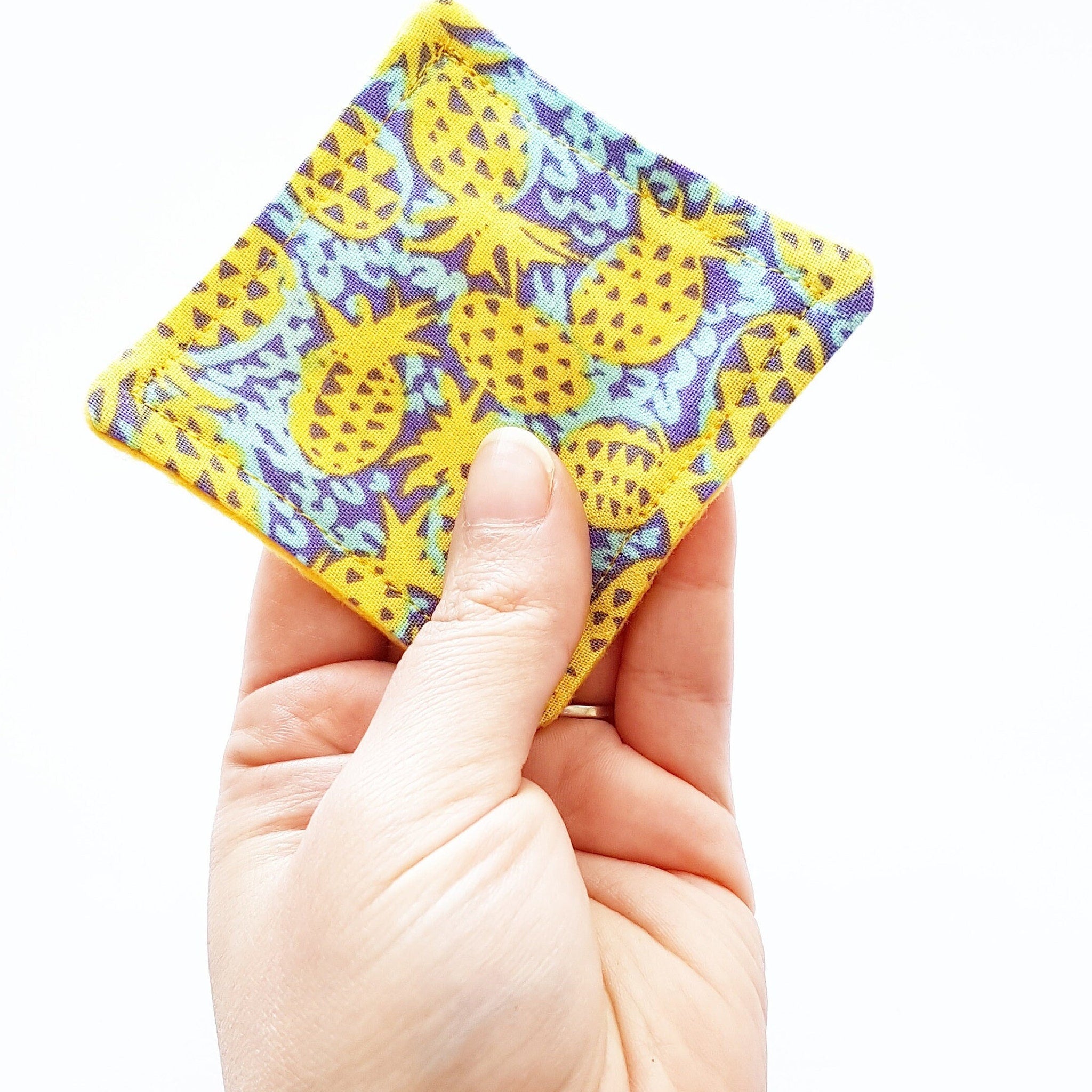 Square fabric with yellow, purple, and blue pineapple pattern held in hand against white background