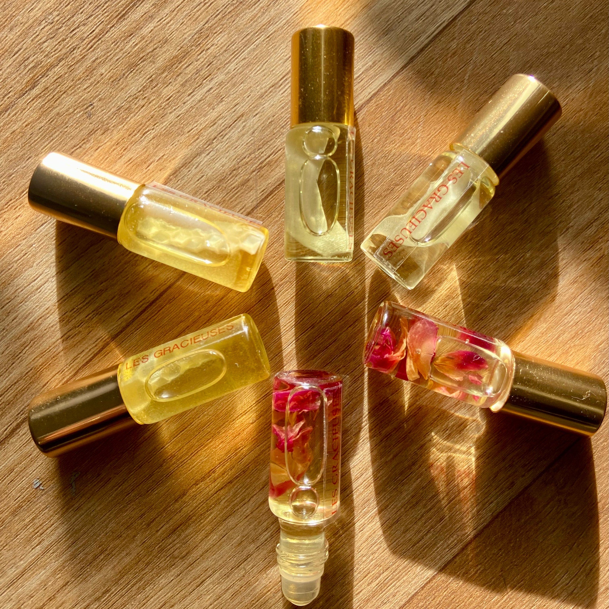 Perfume bottles with gold caps, clear glass, and floral inclusions, on wood surface, LES GRACIEUSES text visible
