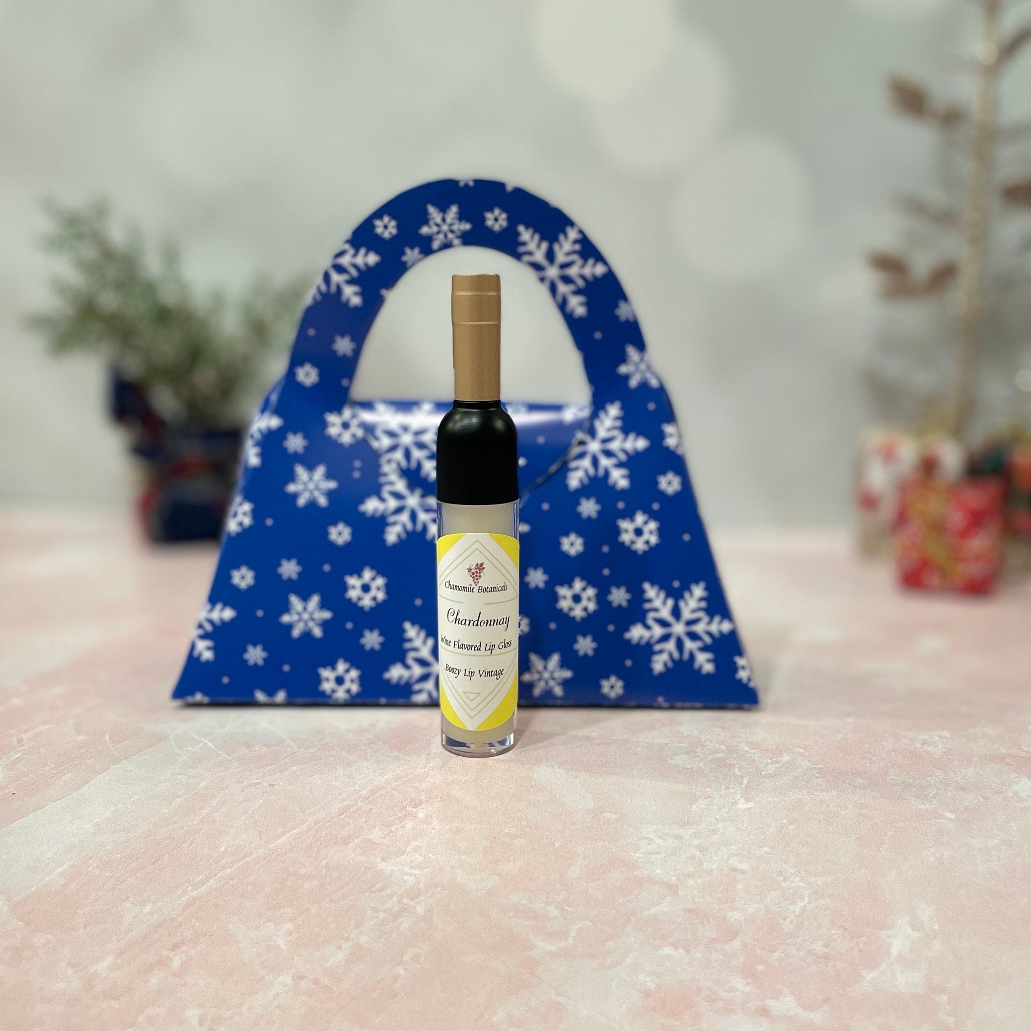 Lip gloss bottle with black, yellow label, and gold cap, in front of a blue snowflake gift bag
