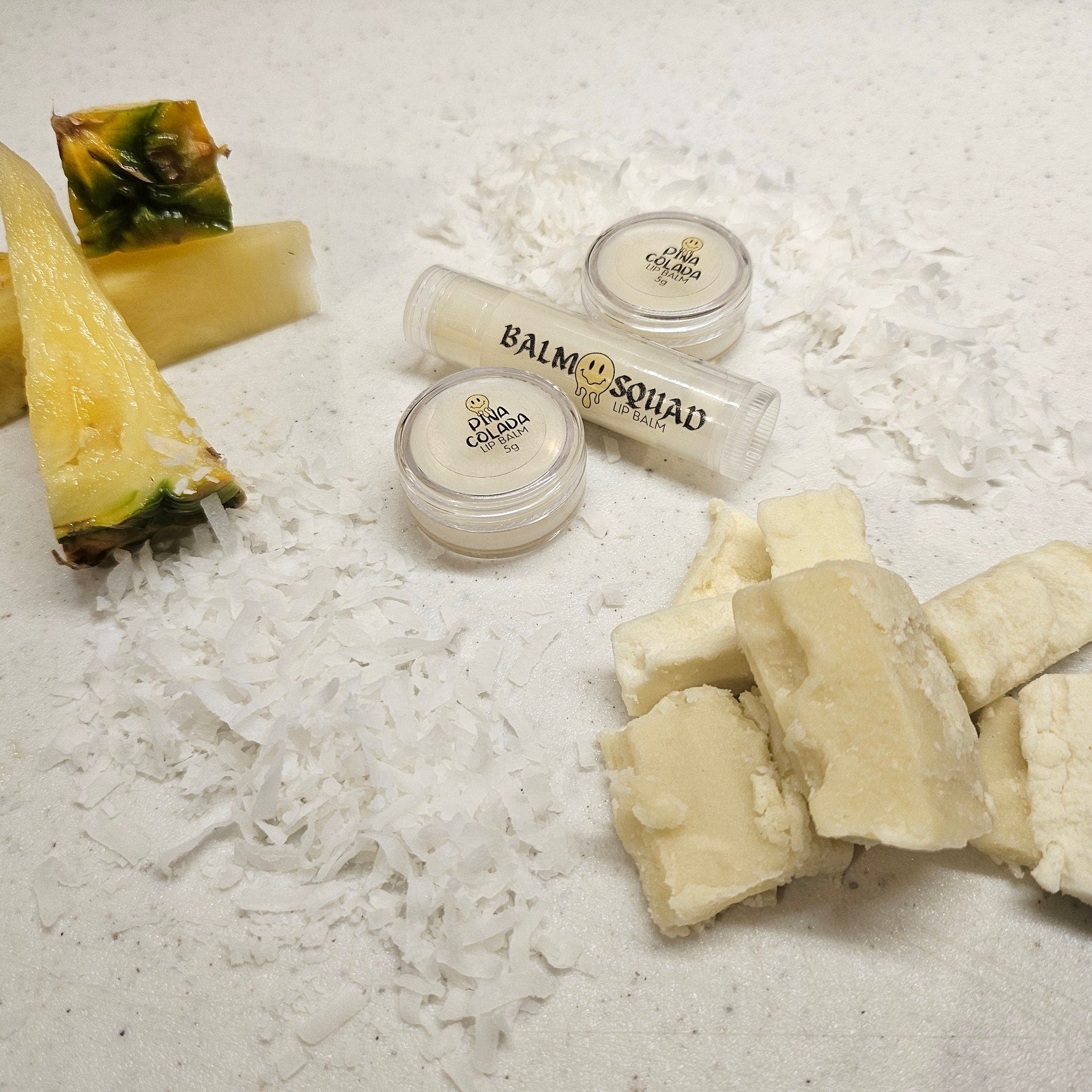 Lip balm tubes and jars with white and yellow accents, surrounded by coconut flakes and fruit, Balm Squad brand
