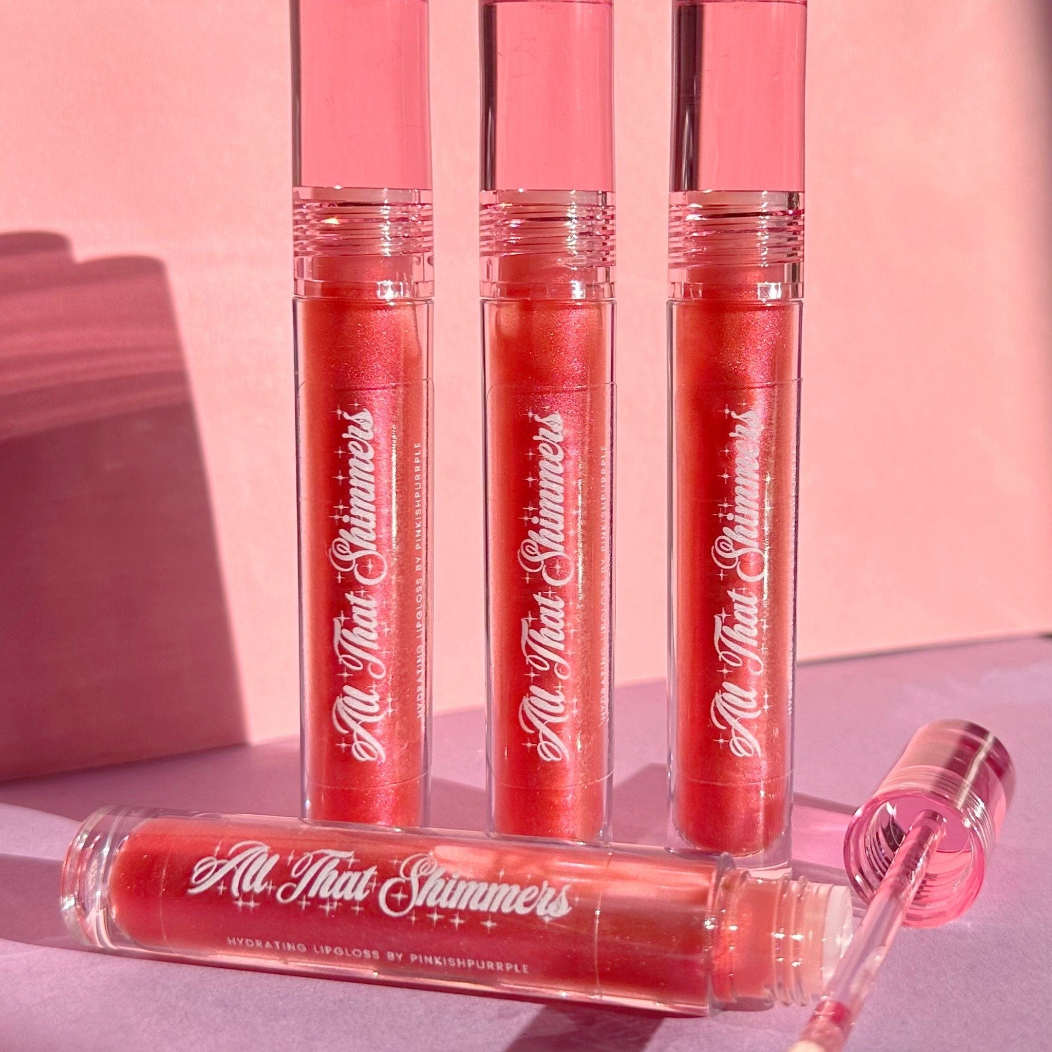Lip gloss tubes in pink, clear, and red with "All That Shimmers" text, one open with applicator visible