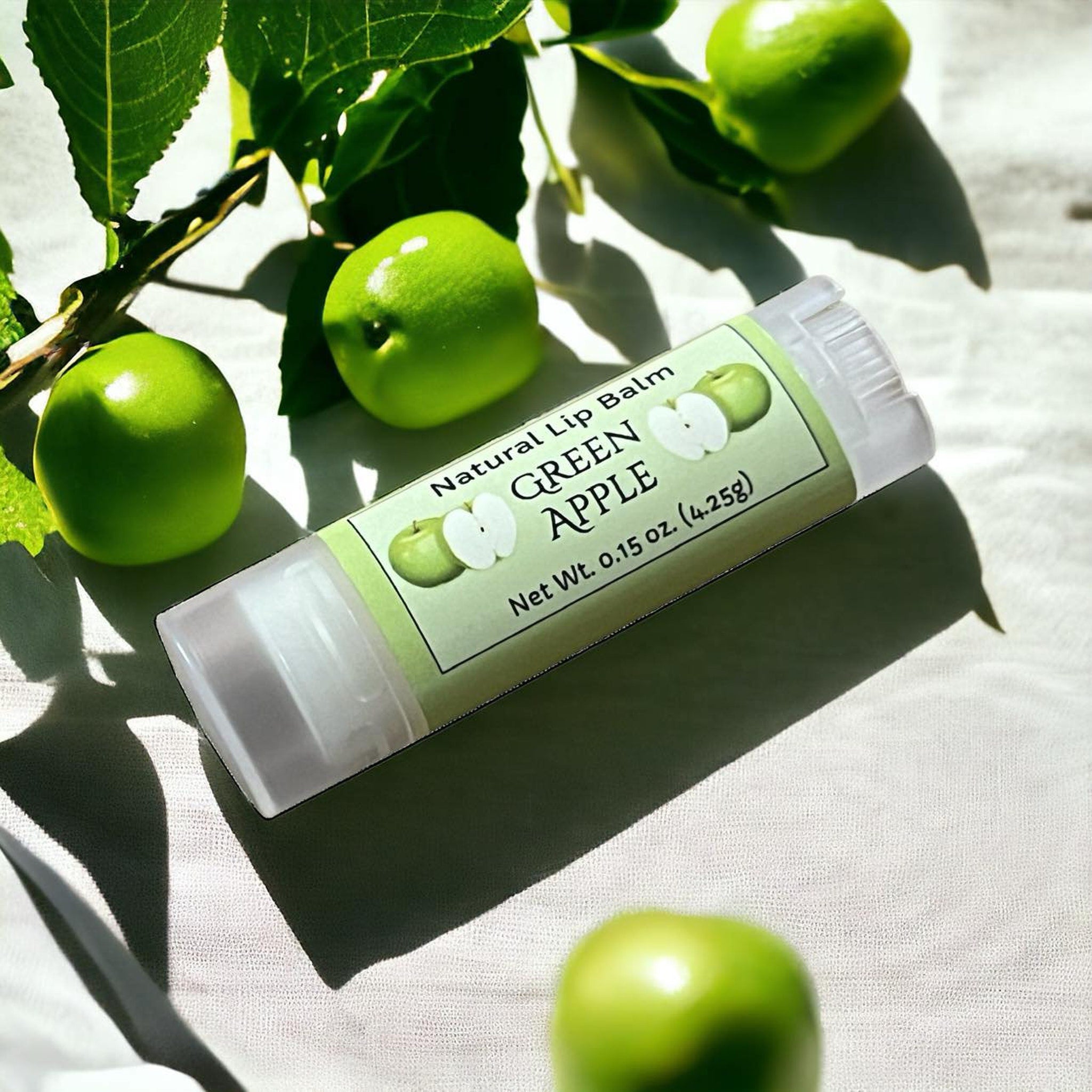 Lip balm tube with green apple label, clear plastic, and green apples on white fabric