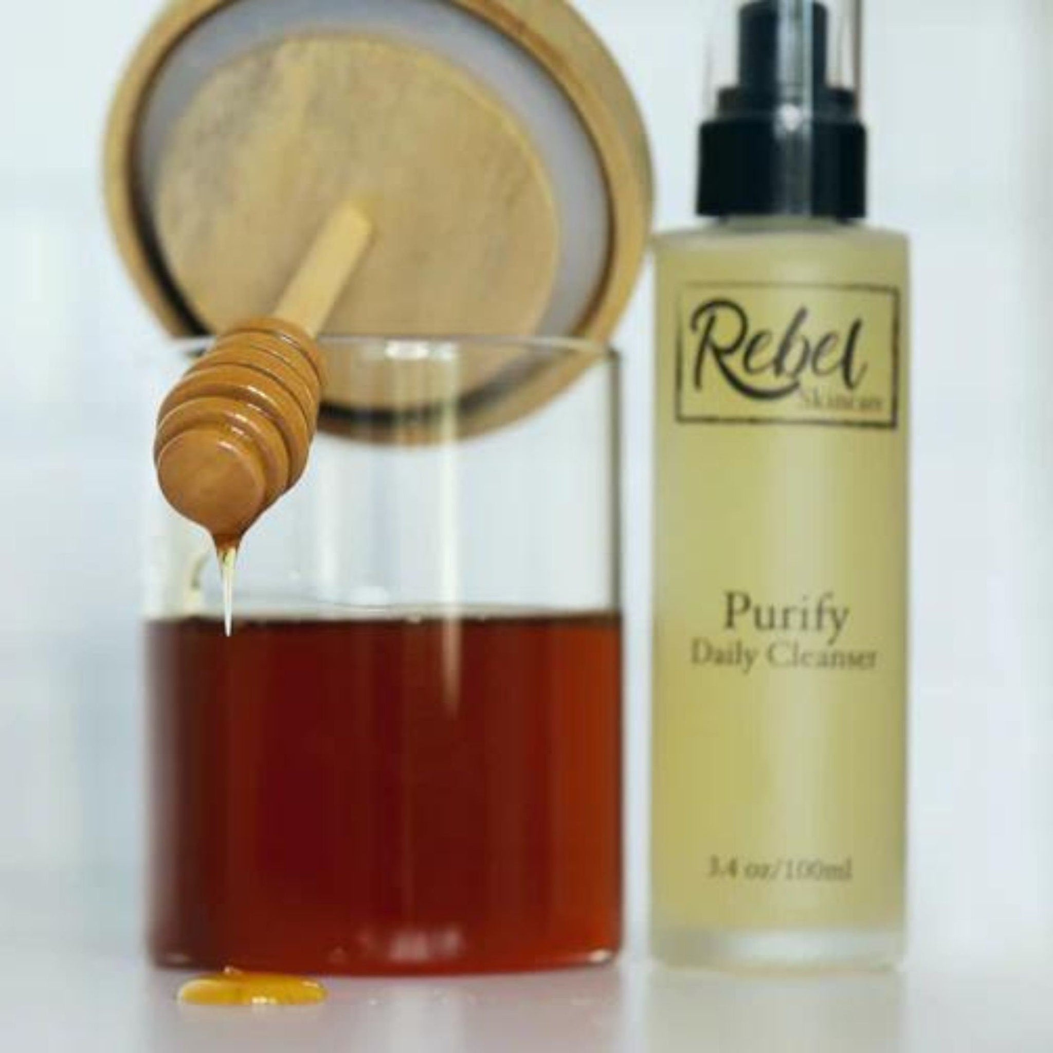 Honey dipper with honey dripping, glass jar with honey, and Rebel Skincare Purify bottle on white surface