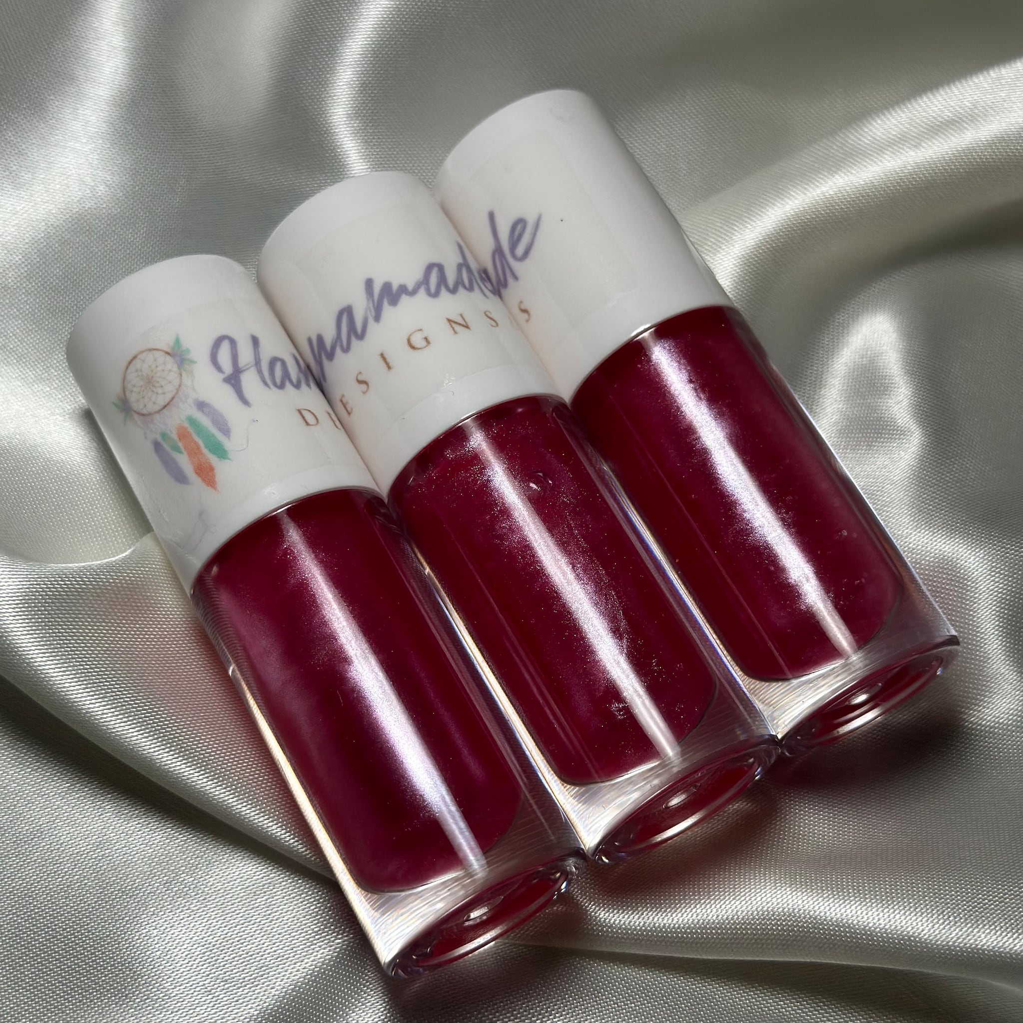 Lip gloss tubes with red gloss, white caps, and "Haypamade Designs" text on satin fabric background